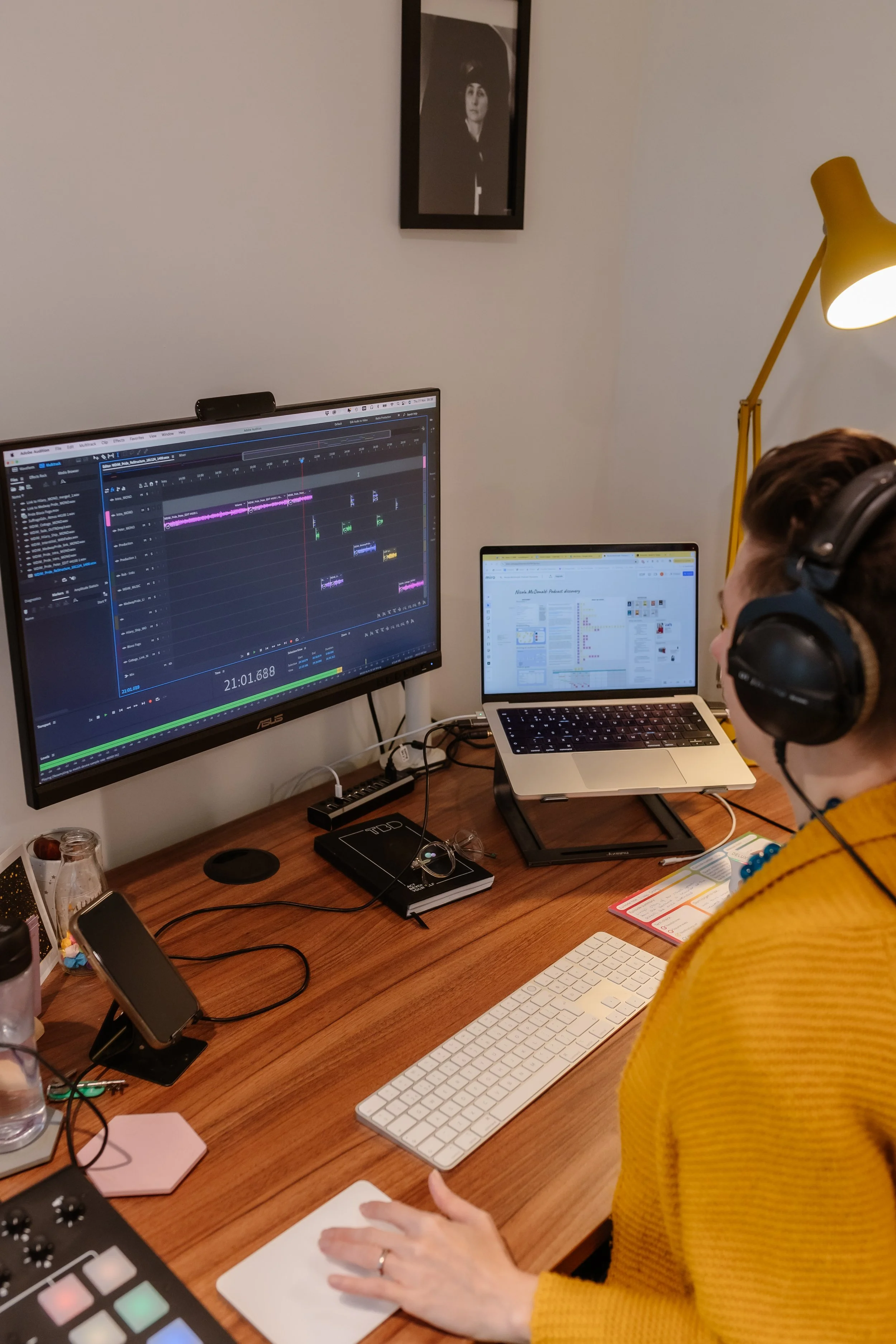 Suze is pictured sitting at her desk. The shot is taken over her left shoulder. She is wearing headphones and looking at her computer screen which shows an Adobe Audition session. She has her lap top open to the right of her screen.