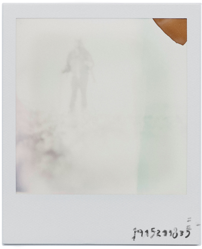 Faded Polaroid image with a ghostly silhouette of a person standing, slightly blurred. The top right corner has a brown discoloration. Mysterious and nostalgic tone.