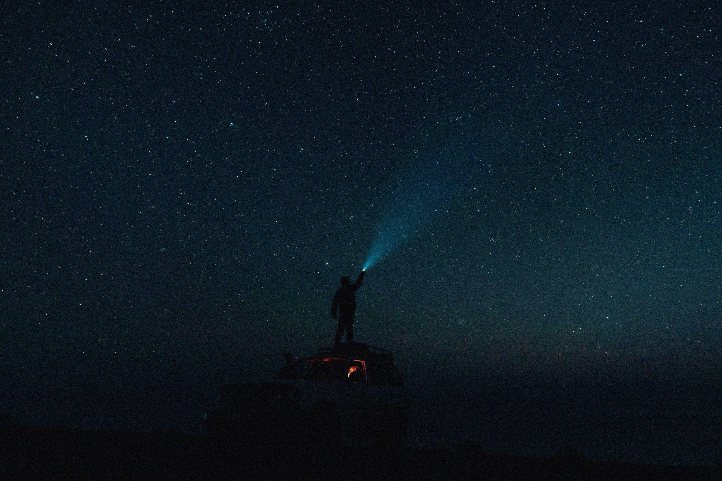 Silhouette of a person standing on a car roof under a starry night sky. They shine a flashlight upward, creating a sense of wonder and exploration.