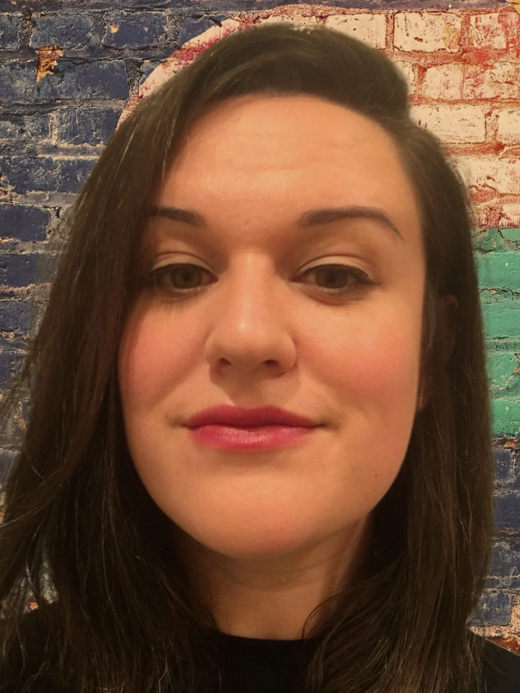 An artistic snapshot of How it Ends Studio co-founder Stephanie Resendes. A woman with dark brown hair and green eyes smiling into the camera against a brightly colored brick wall.