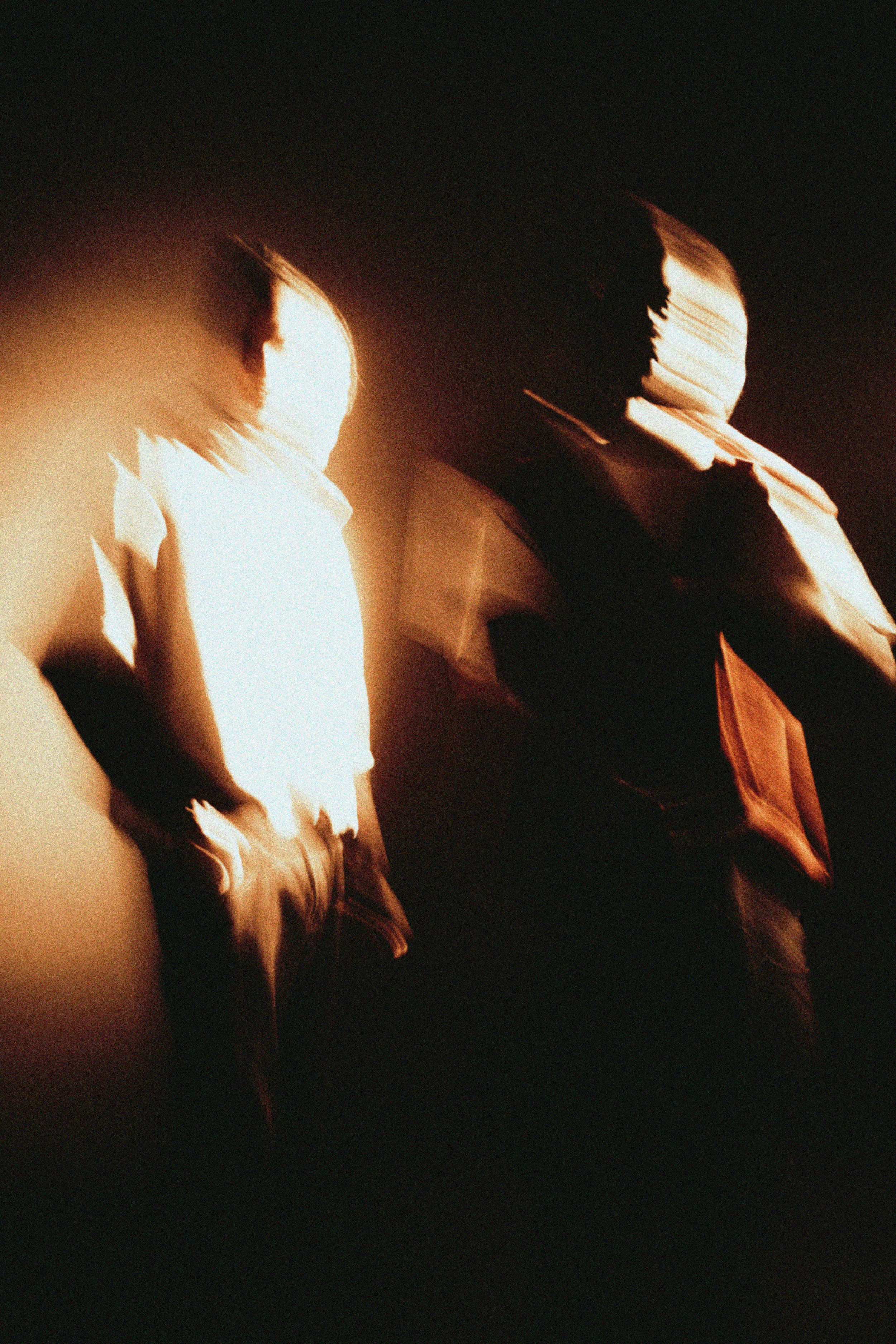 Blurred figures in warm light create an abstract, dream-like atmosphere. The motion and rich tones evoke mystery and emotion against a dark background.