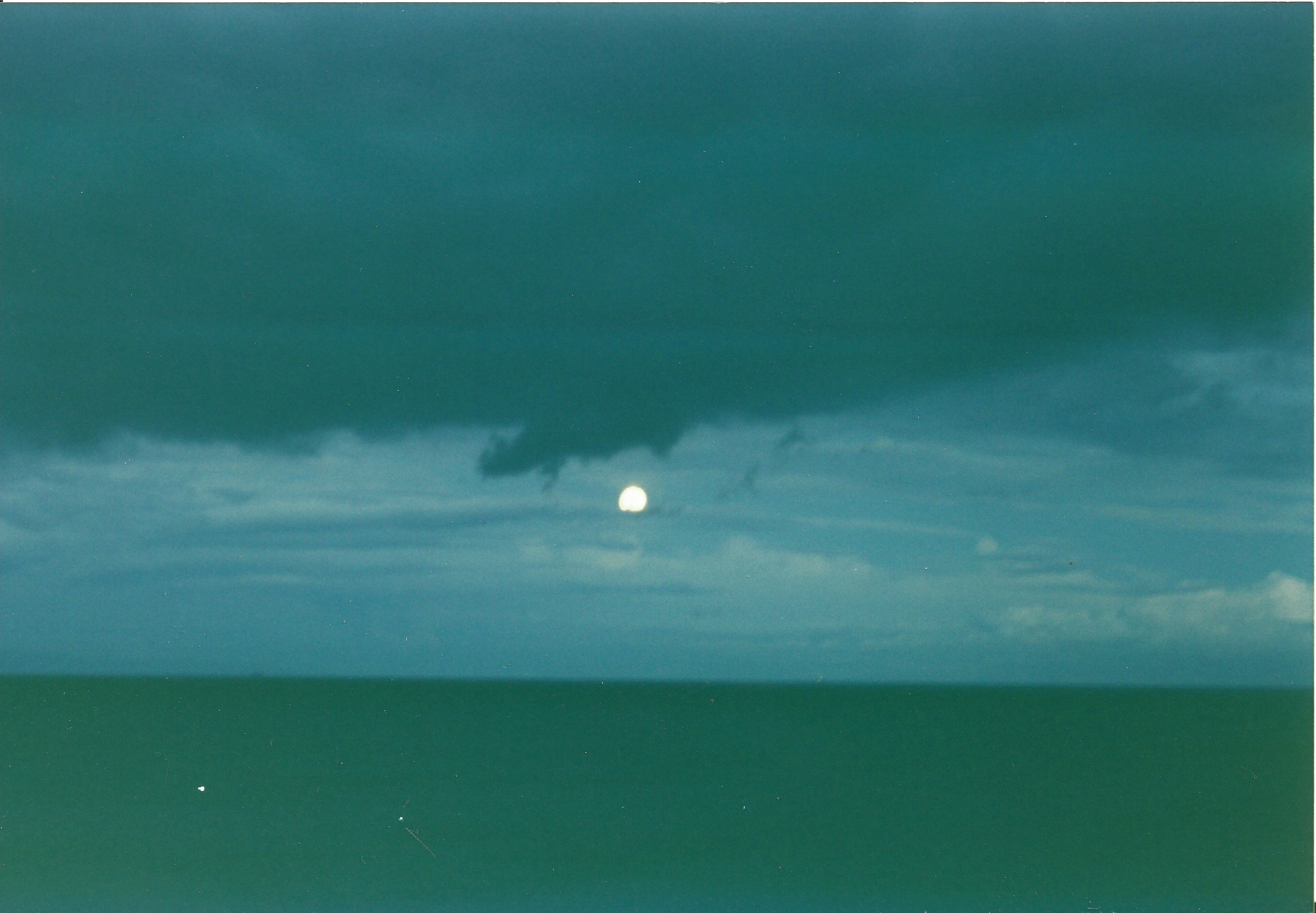 A full moon glows dimly behind dark clouds over a vast, calm ocean, creating a serene and mysterious nighttime seascape.