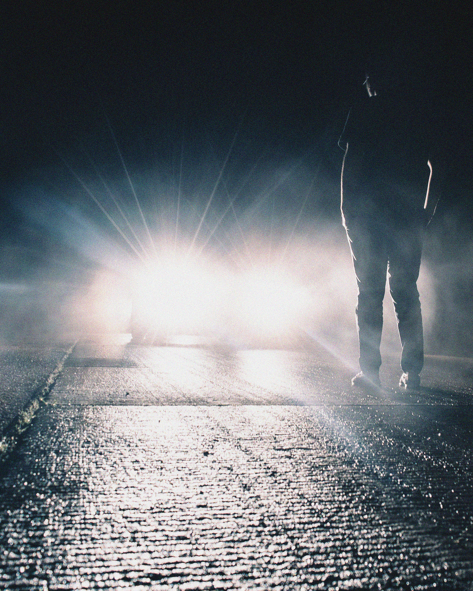 A lone figure stands on a misty road at night, silhouetted against bright car headlights. The atmosphere is tense and mysterious, with shadows and fog.