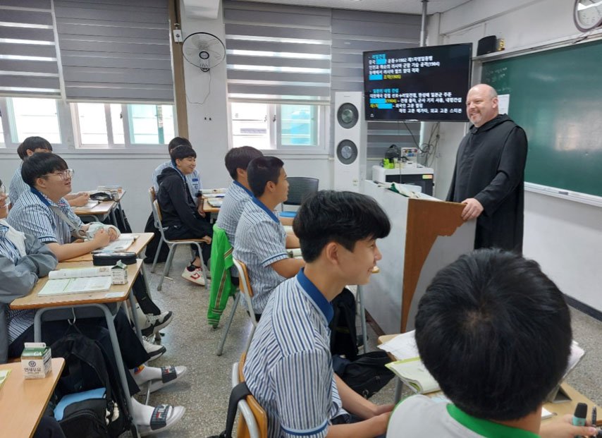 A Catholic priest in black robes stands in front of a classroom with students engaged in conversation.