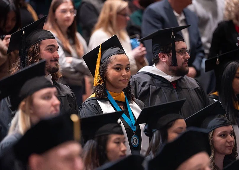 A group of graduates in caps and gowns, with one graduate in the foreground wearing a medal, standing out among the crowd.
