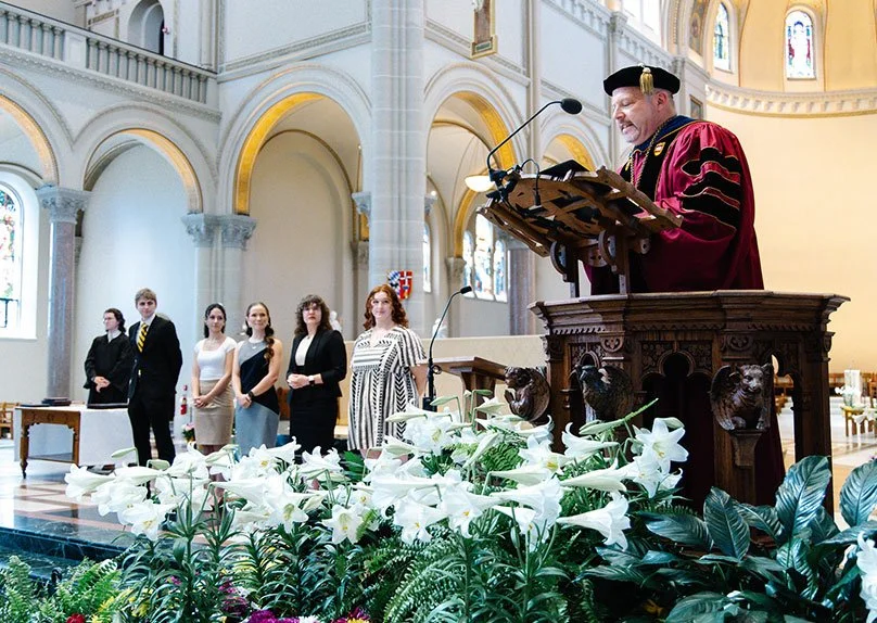 Father Paul in academic regalia delivers a speech at a ceremony, with students standing on stage in the background.