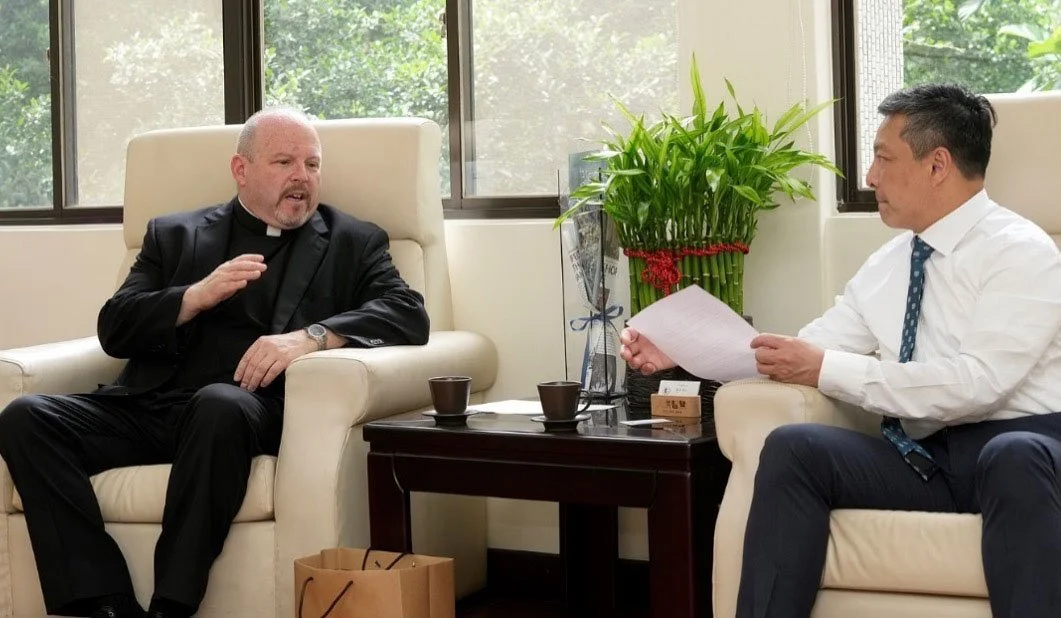 Two men sit in a lounge area engaged in a discussion, the man on the left is dressed in black wearing a white priest’s collar and the man on the right is wearing a white shirt, blue pants, and printed tie.