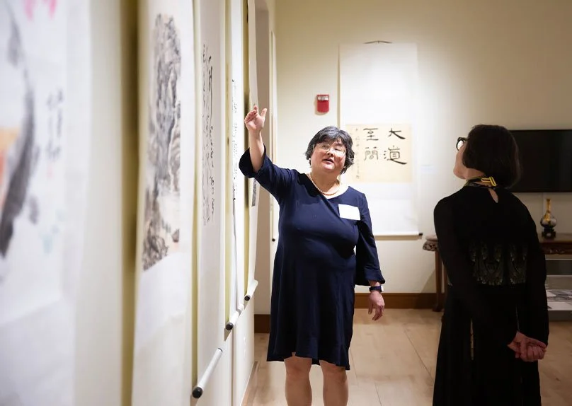 A woman gestures while explaining artwork in a gallery, with a woman listening attentively in the background.