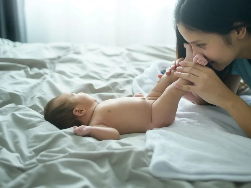 A woman playfully holding a baby's feet while lying on a bed with white sheets, smiling at the baby.