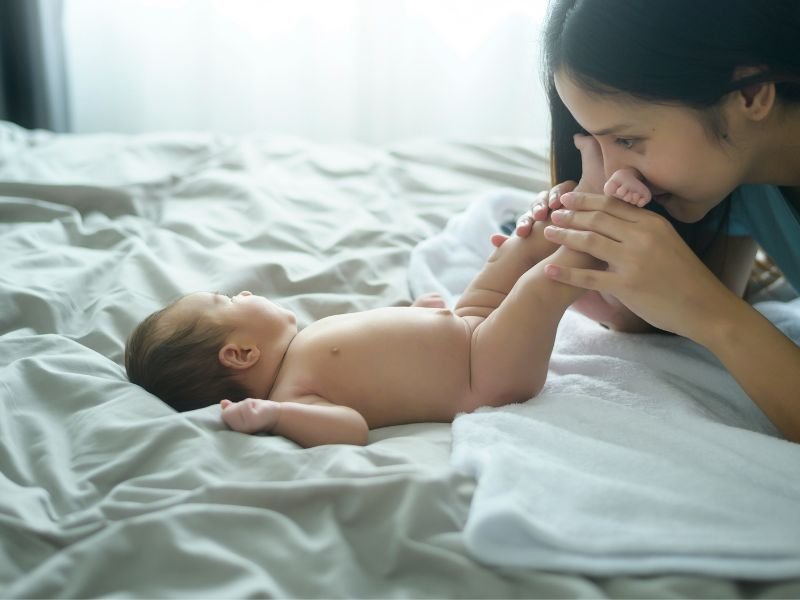 A mother playing with her naked baby on a bed, holding the baby's feet and smiling at each other in a softly lit room.