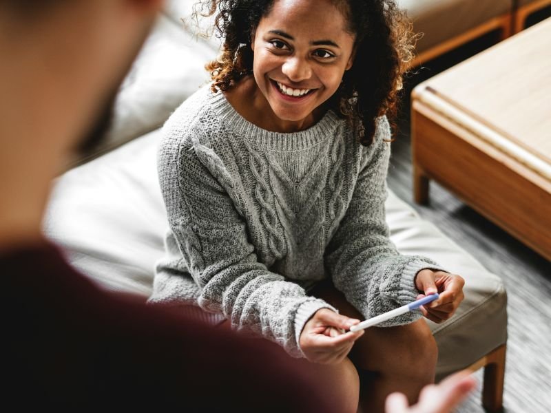 Young woman with curly hair smiling and holding a pregnancy test while talking to a person whose back is to the camera.