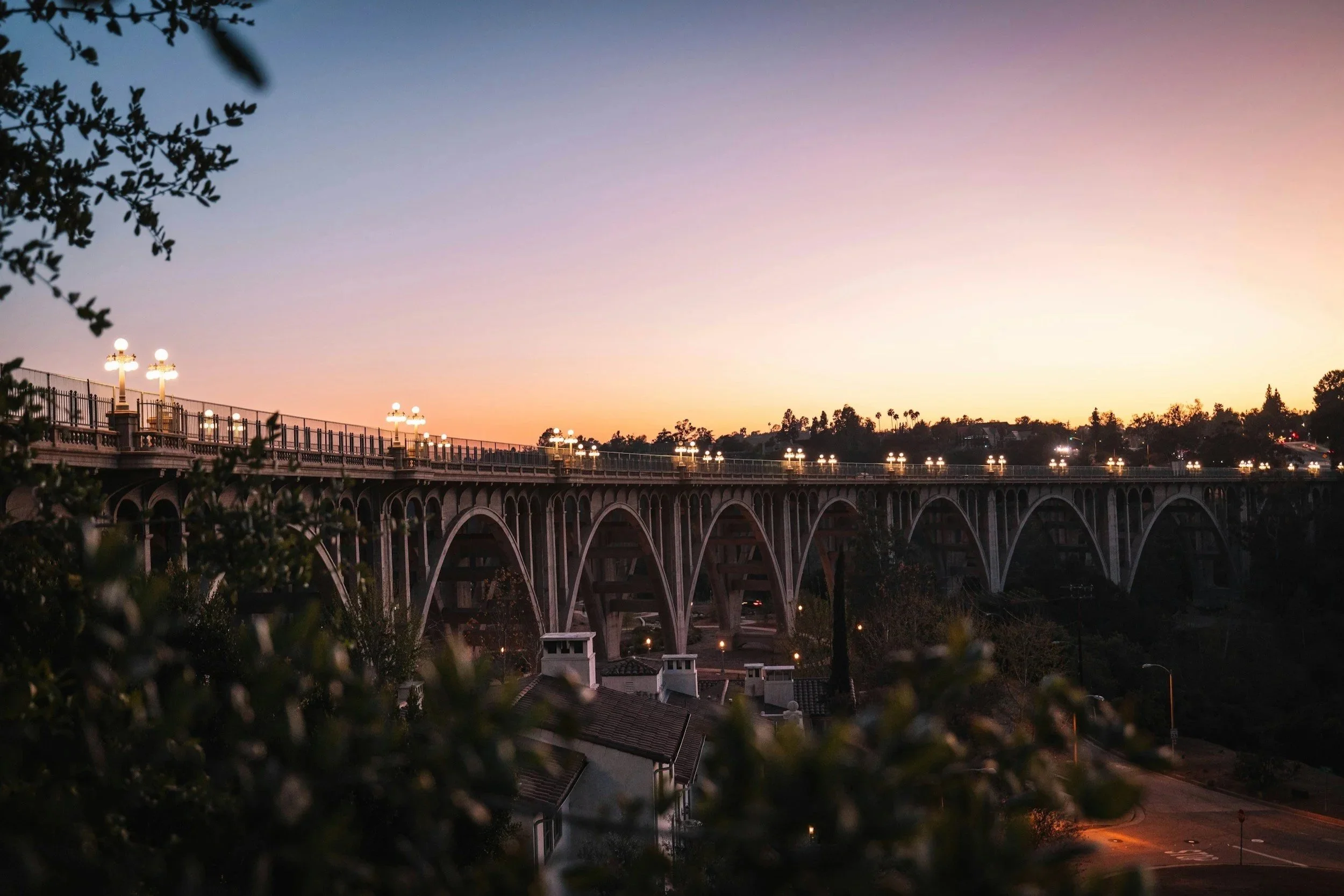 The Colorado Street bridge in Pasadena with multiple arches and streetlights spans over a darker area, with a sunset sky in the background showing pink, orange, and purple hues. Trees and houses are visible below and on the horizon.