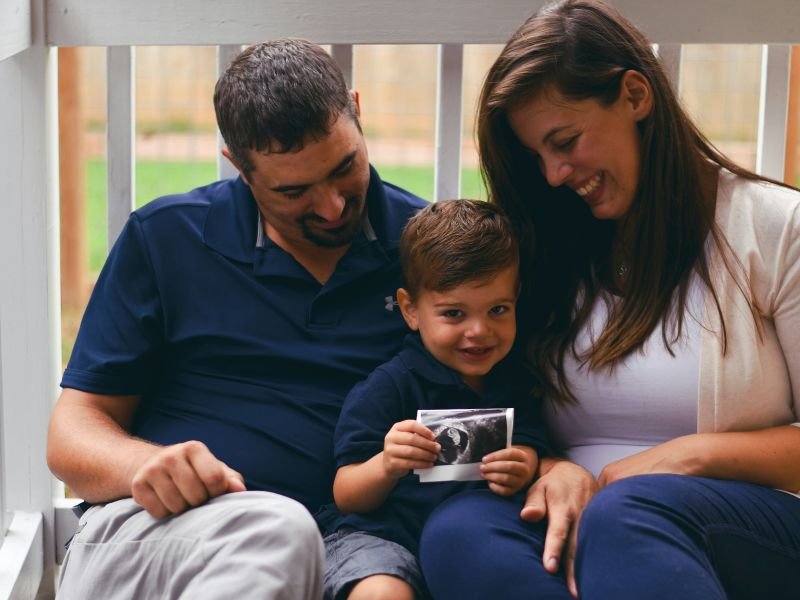 A family of three sitting outdoors on a porch, with a man, a woman, and a young boy holding an ultrasound picture, all smiling and looking happy.