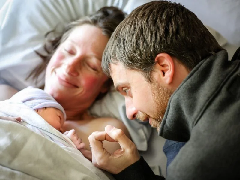 A mother in a hospital bed with her newborn baby, father lying beside her, reaching out and touching the baby's hand, all three sharing a tender moment.