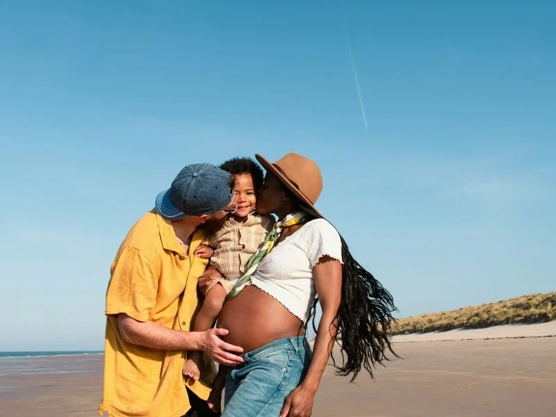 A family enjoying a moment at the beach, with a pregnant woman and a young boy, sharing a kiss.
