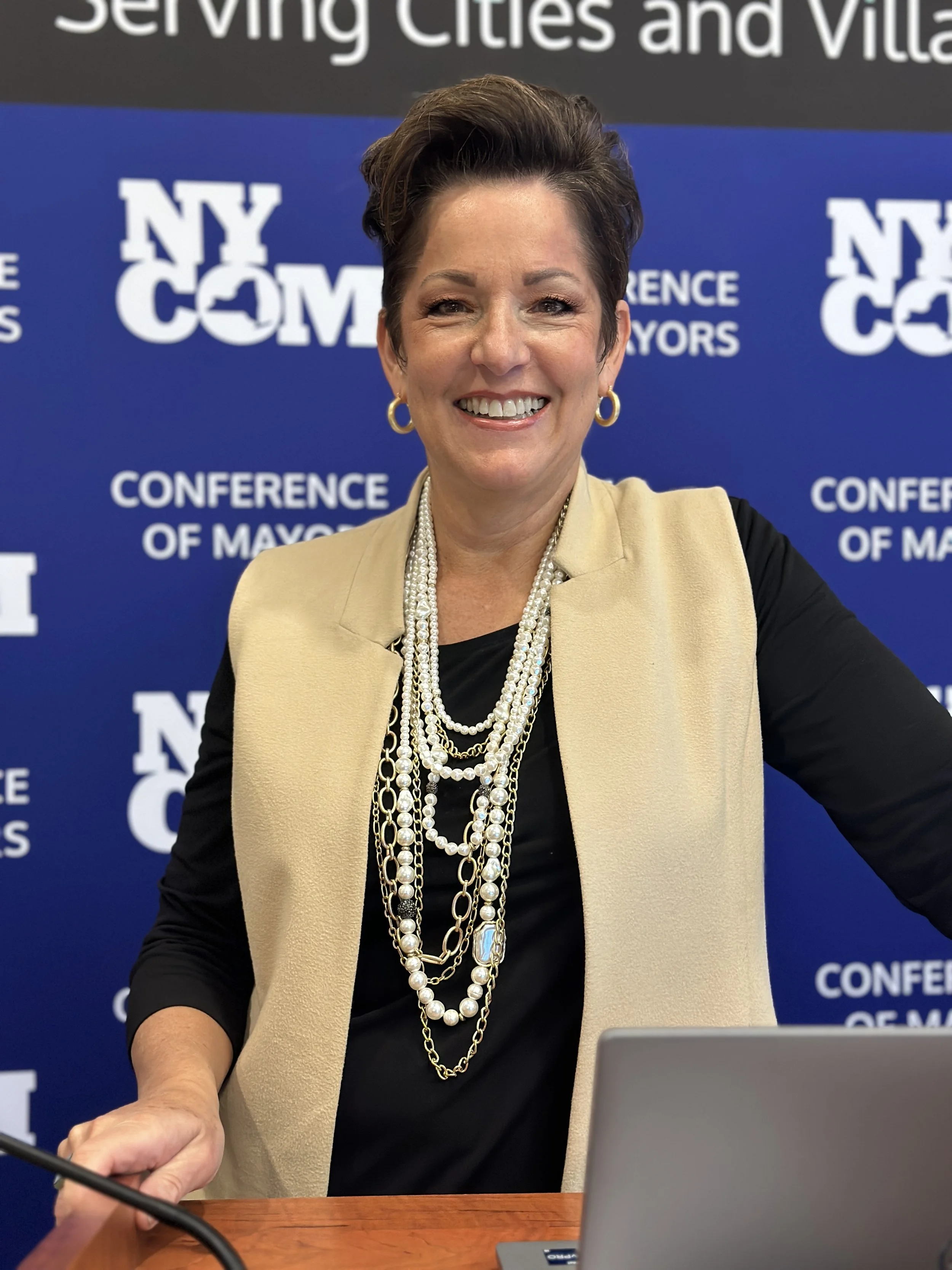 A woman smiling at a conference, wearing a beige vest, black top, and multiple pearl and gold necklaces. She is standing in front of a blue background with the text 'NY COM' and 'CONFERENCE OF MAYORS'.