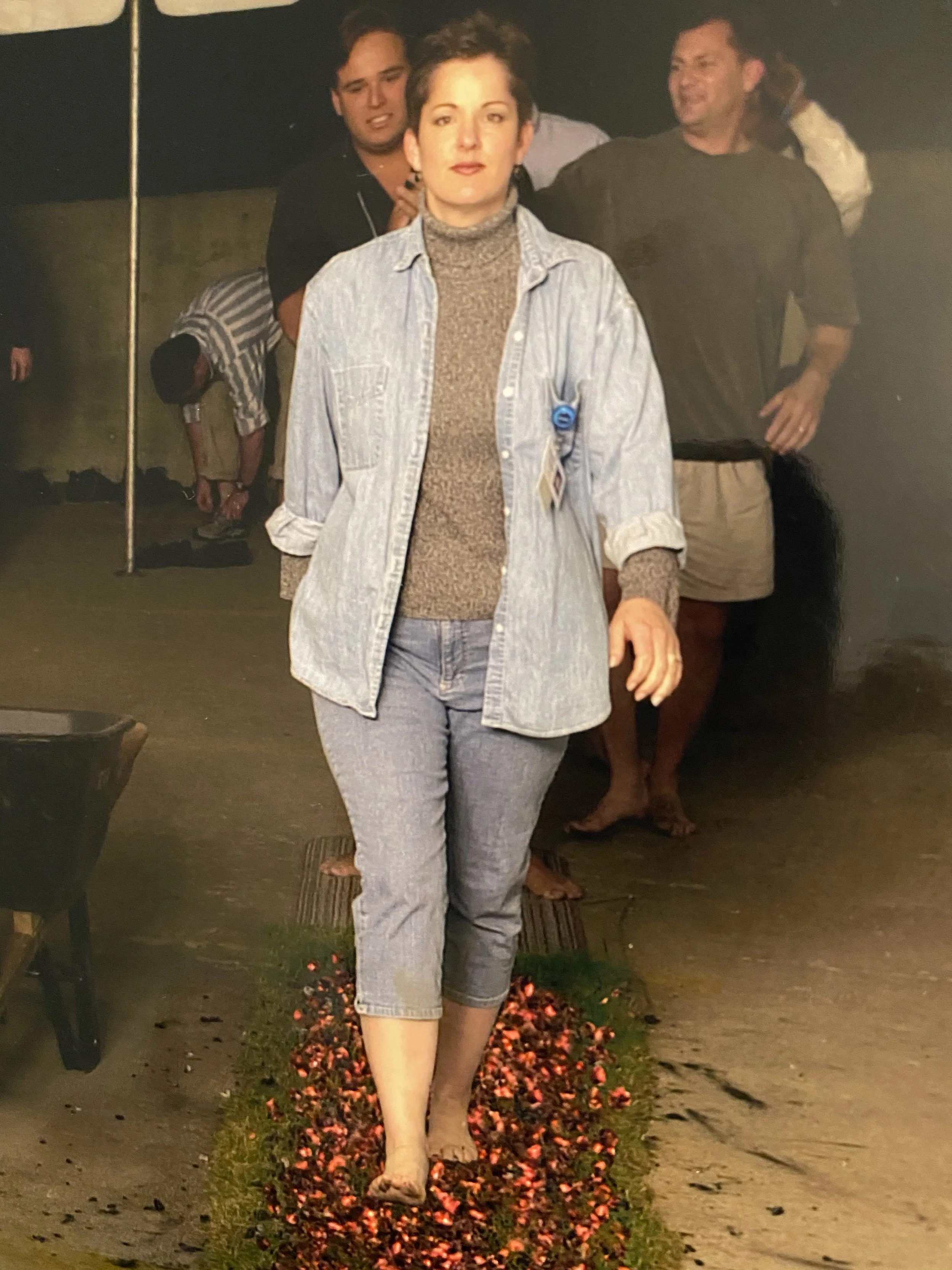 Woman standing on a grassy path with flower petals, wearing jeans, a turtleneck, a denim jacket, and beige shoes, with three people behind her, at night outdoors.