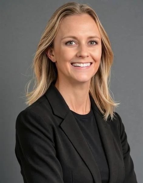 A professional woman with blonde hair smiling, wearing a black blazer and black shirt against a gray background.