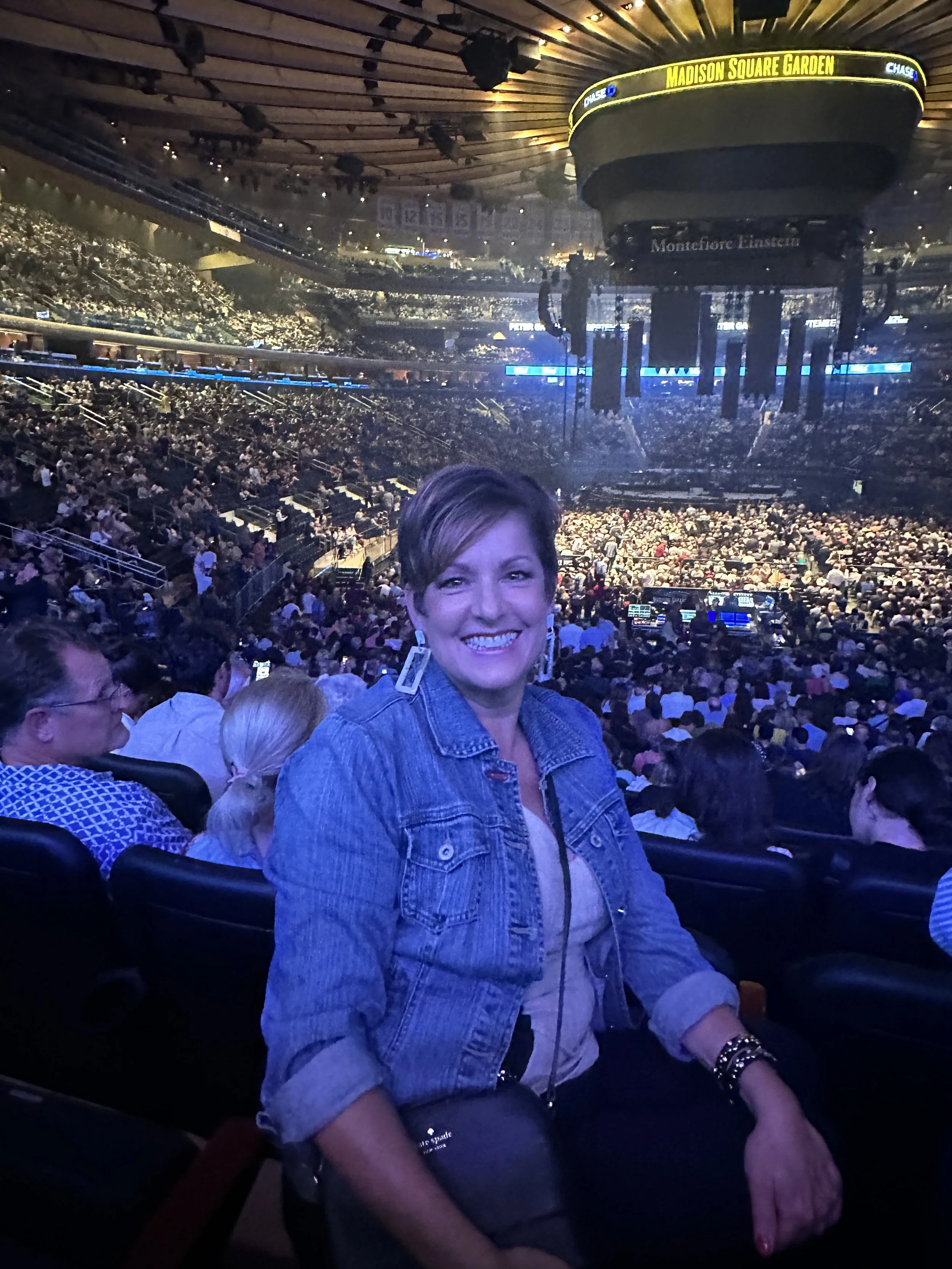 Smiling woman in denim jacket sitting in a crowded arena at Madison Square Garden with the stage and audience visible in the background.