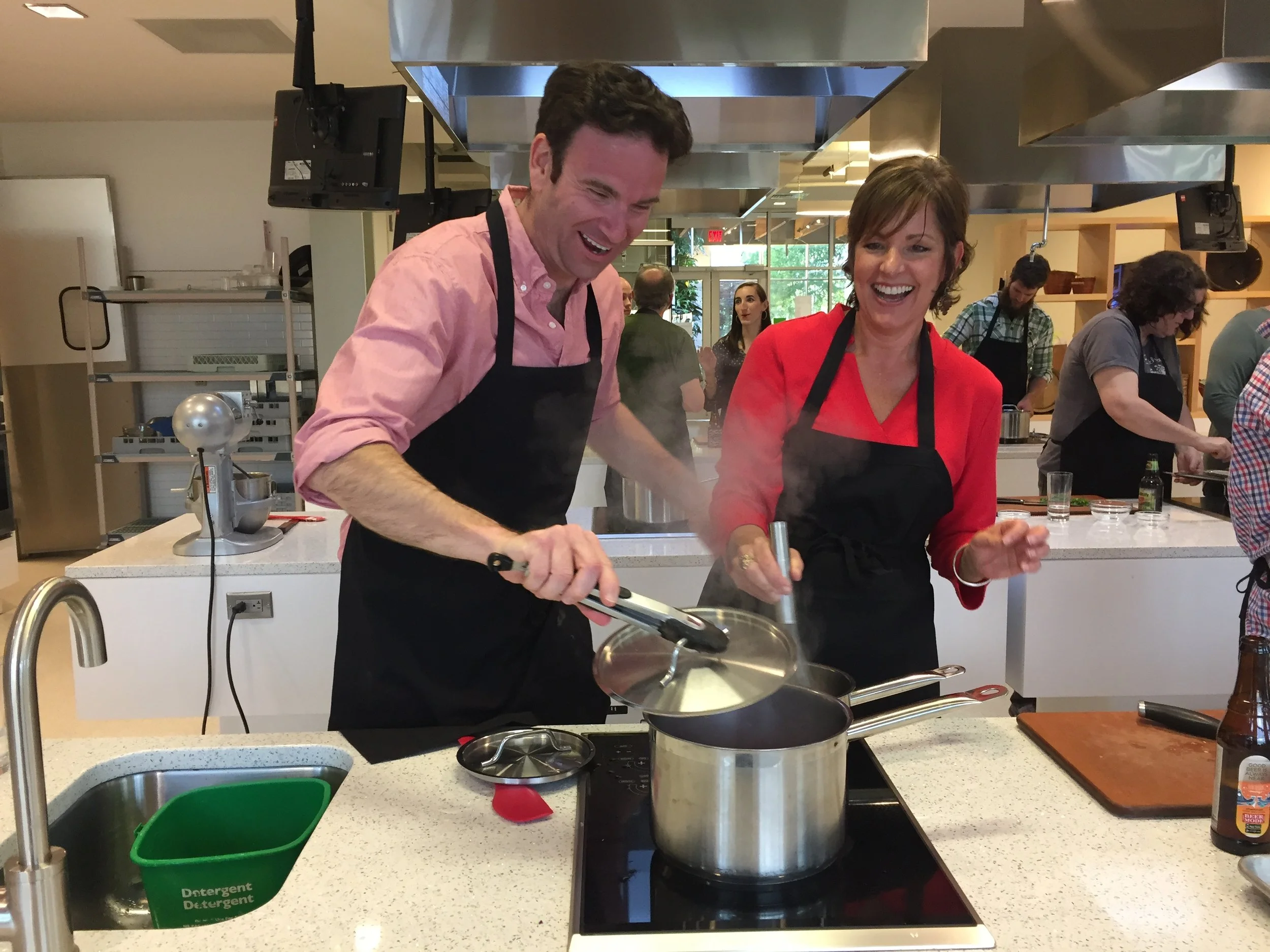 Two people cooking and laughing in a kitchen studio during a cooking class, wearing black aprons, with multiple other people cooking in the background.
