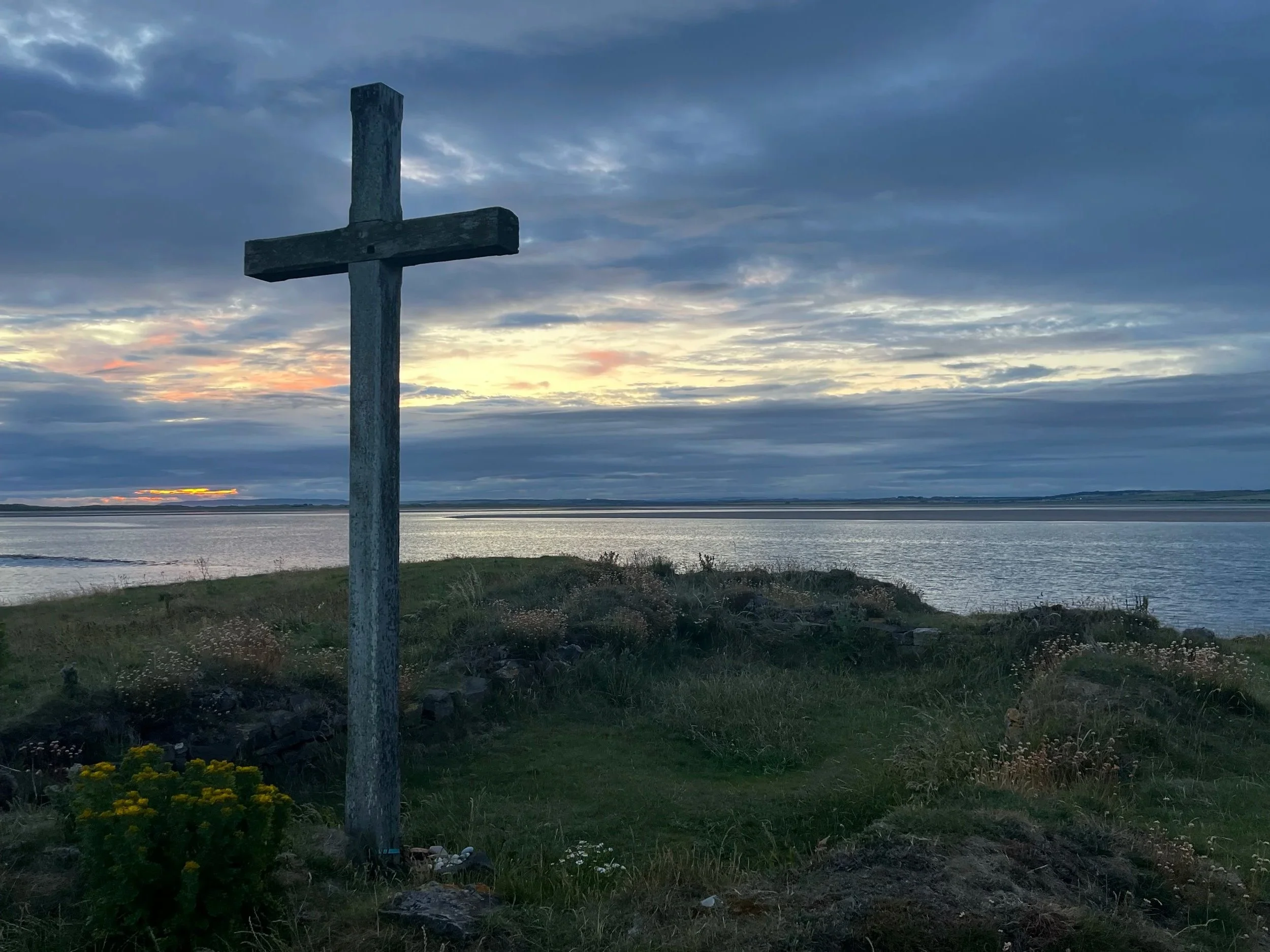 A large wooden cross stands on a grassy shoreline overlooking a body of water during sunset with a cloudy sky.