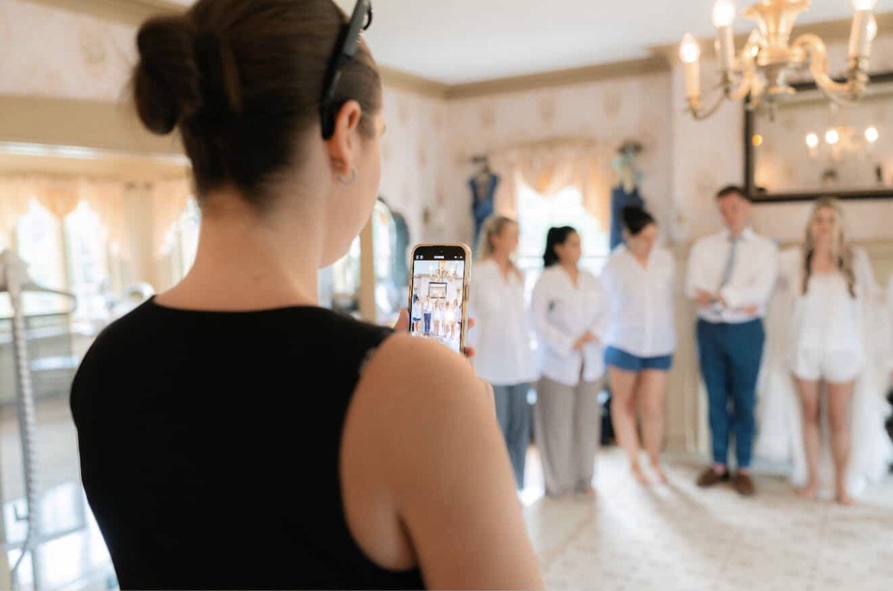 Woman taking a photo of a group of people standing inside a house, with some of them dressed in white and blouses, near a chandelier.