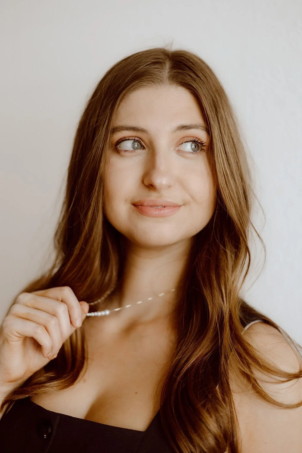 A young woman with long, wavy auburn hair holding a pearl necklace, looking to the side with a slight smile, against a plain white background.