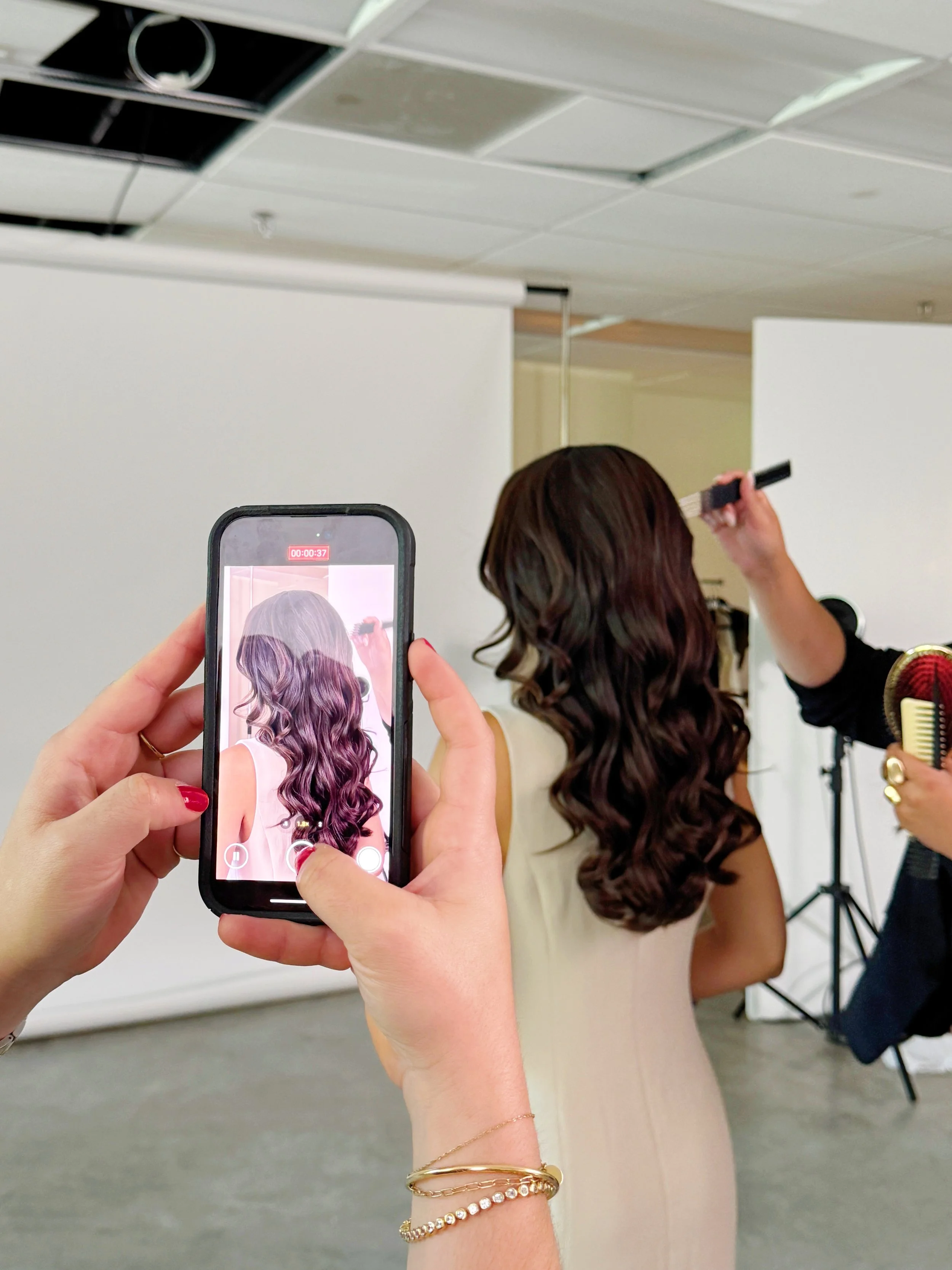 A woman with long, wavy brown hair is having her photo taken in a studio, with a person using a smartphone to capture the image. The woman is wearing a light-colored sleeveless dress, and a person is styling her hair with a flat iron. The studio has a white backdrop and professional lighting equipment.