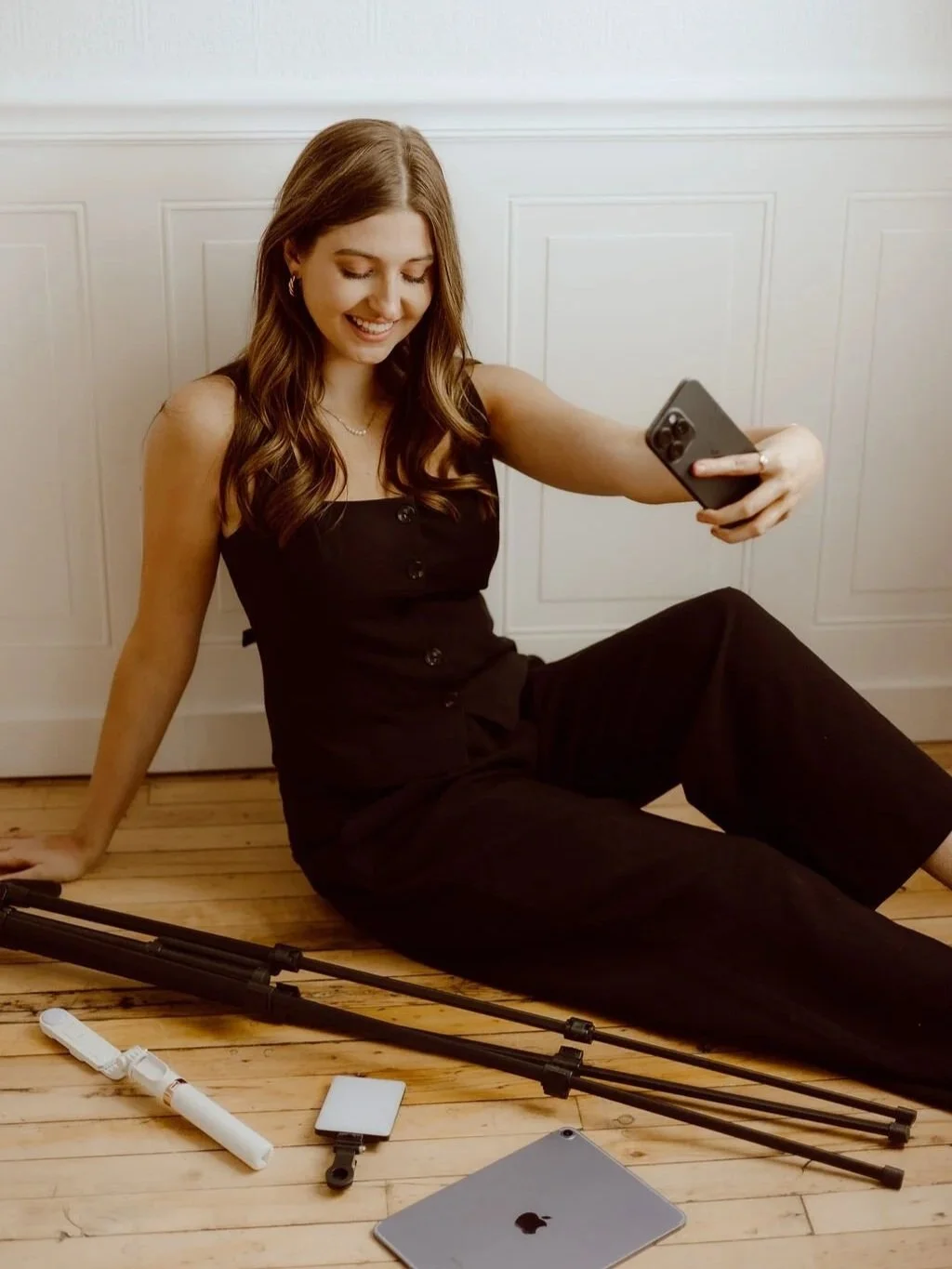 A woman sitting on a wooden floor, smiling and taking a selfie with her smartphone. She has long wavy brown hair, is wearing a black sleeveless jumpsuit, and has photography equipment including tripod and accessories laying nearby.