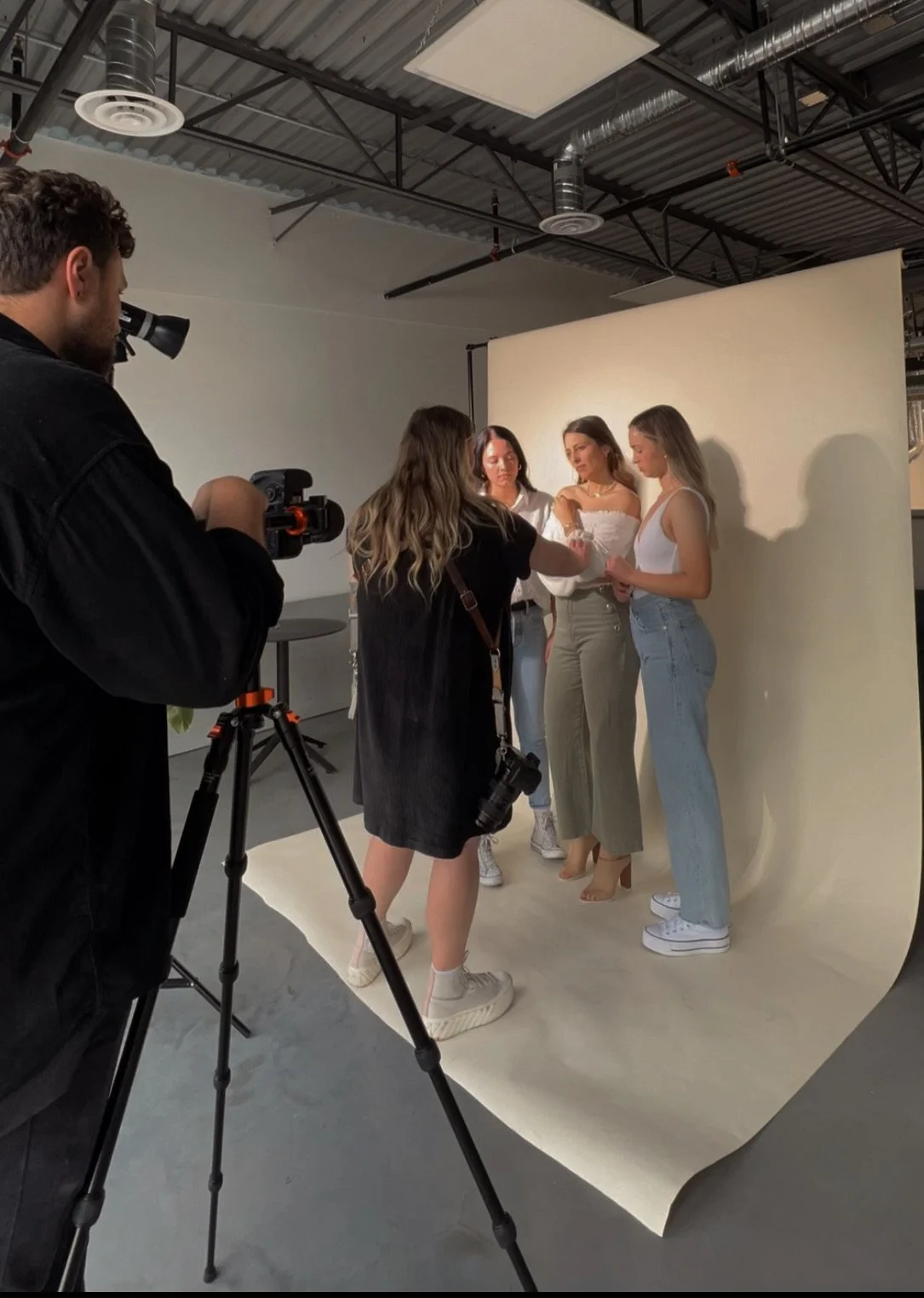 Photoshoot scene with four women standing against a beige backdrop, with a photographer and an assistant adjusting their outfits and poses in a studio.
