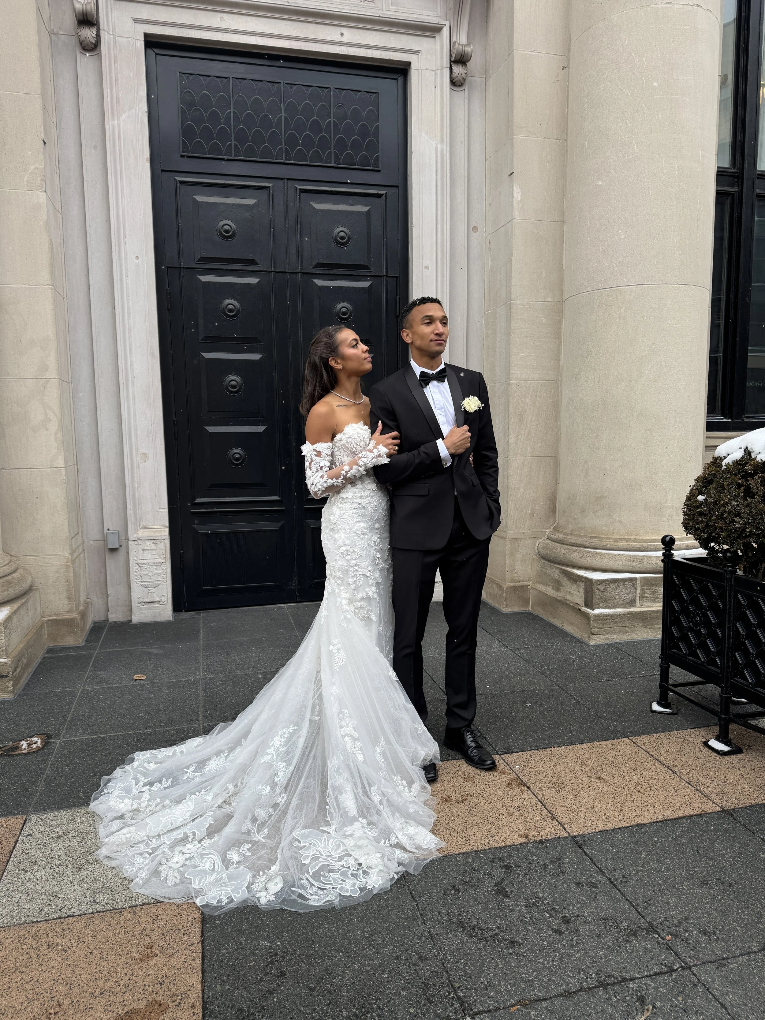 A bride and groom standing outside in front of a large black door on a stone sidewalk. The bride is wearing a white lace wedding gown with off-the-shoulder sleeves and a long train, and the groom is in a black tuxedo with a black bow tie and white shirt. The bride is looking at the groom while holding his arm, and the groom has one hand in his pocket and the other resting on his chest.