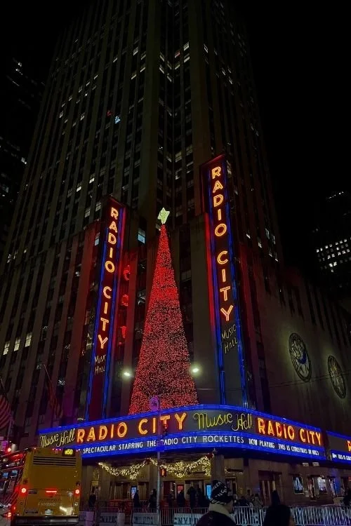 Night view of Radio City Music Hall in New York City decorated with a large Christmas tree with red lights and a star on top, with bright neon signs and people outside.