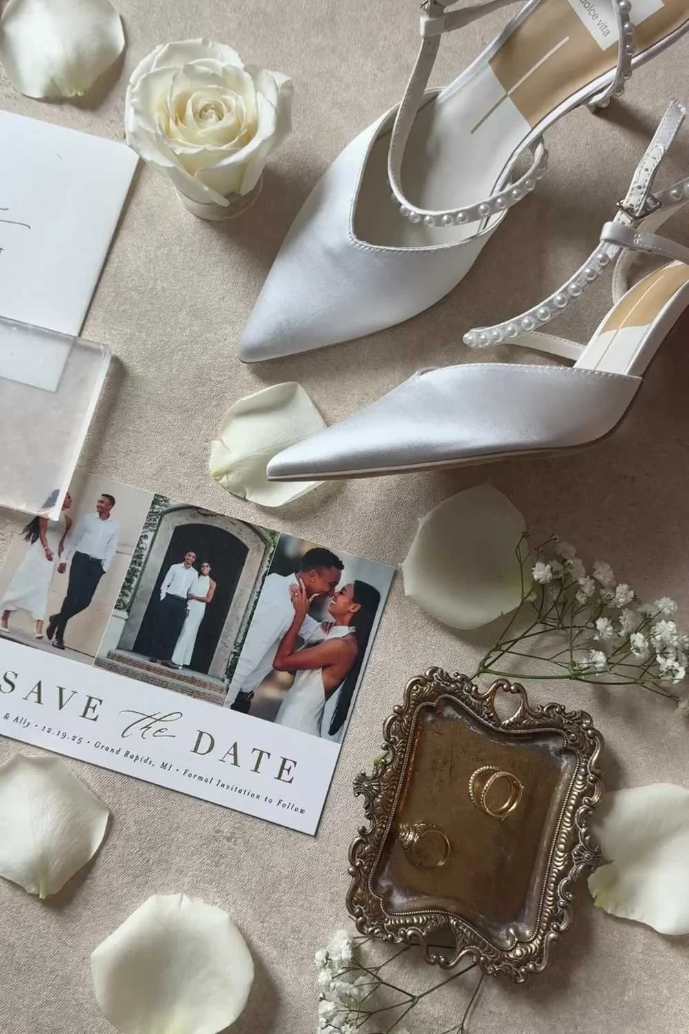 A bride in a white lace wedding dress and a groom in a black tuxedo pose together outside a building with large black double doors. The bride has a long train and the groom is wearing a bow tie and boutonniere.