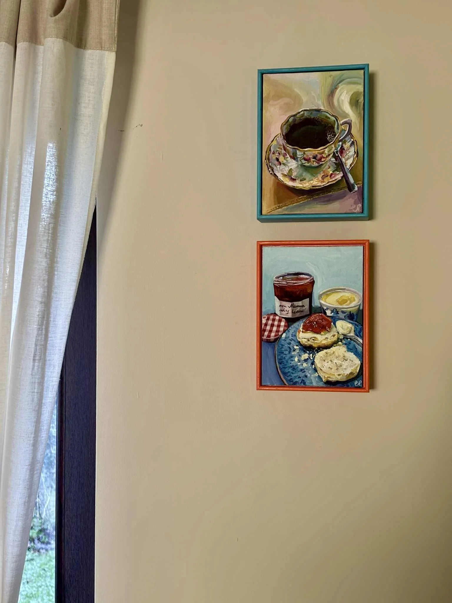 Two colorful paintings of food hung on a beige wall next to a window with curtains.