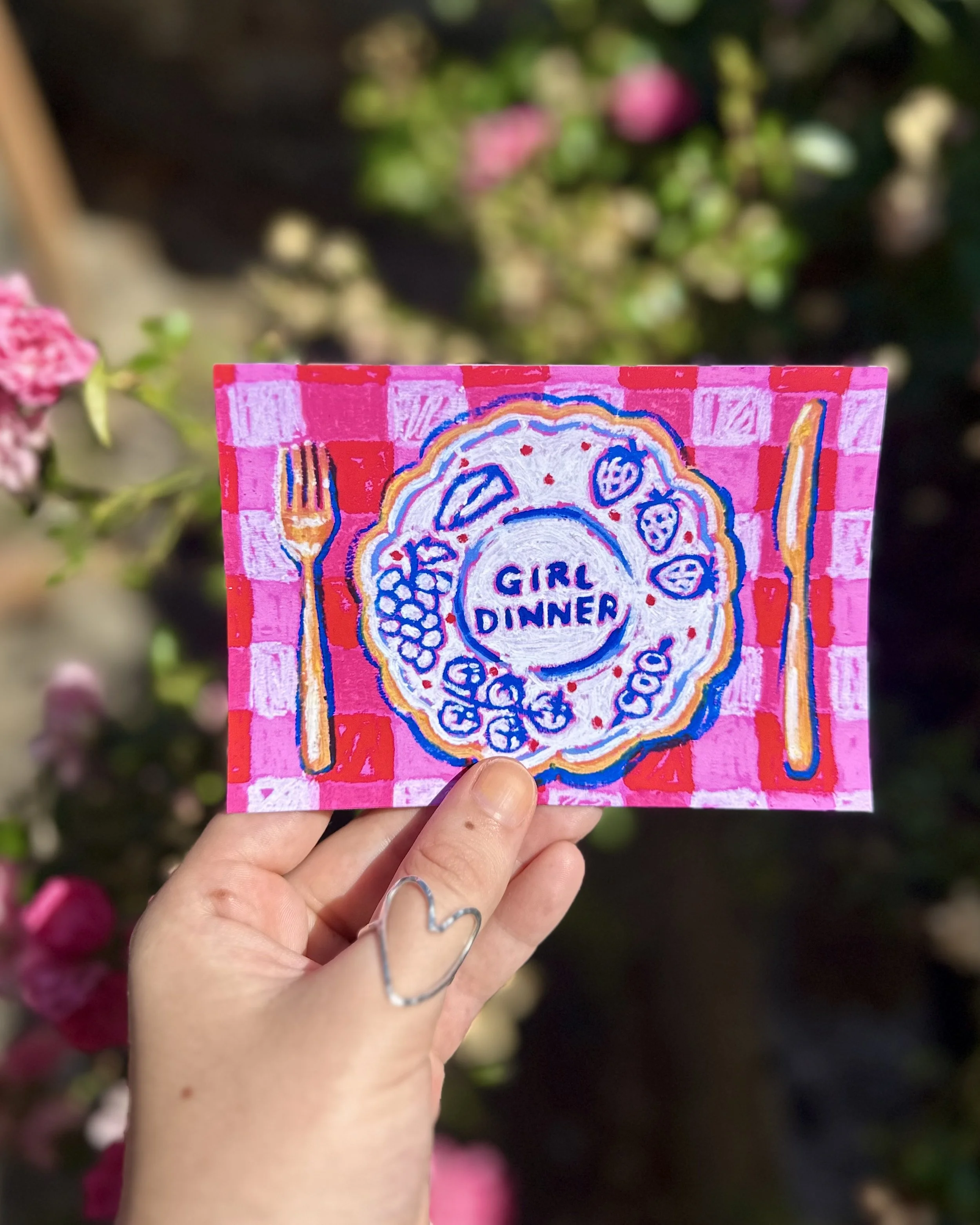 A person's hand holding a colorful card with the text 'Girl Dinner' and a plate illustration, with a blurred background of pink and white flowers and green foliage.