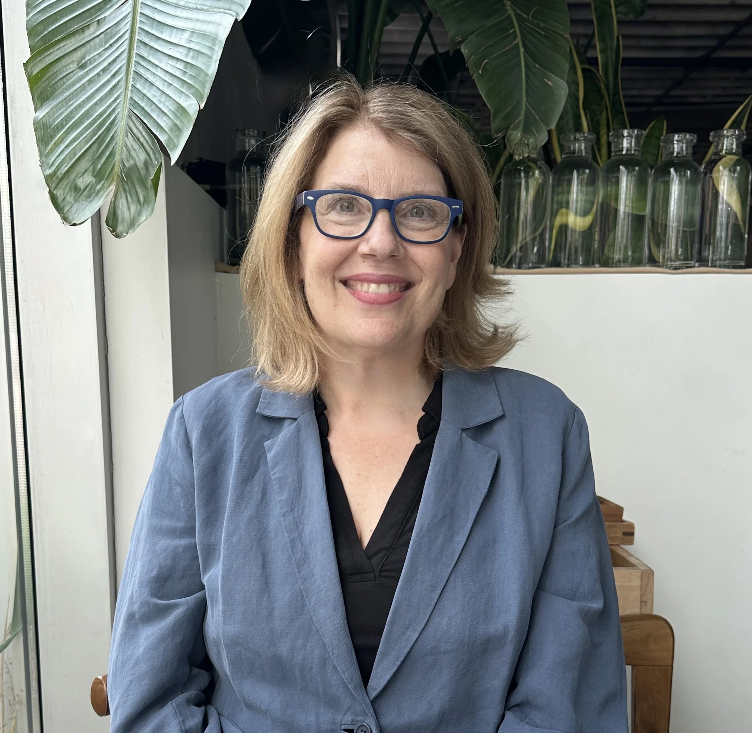 Sara is sitting on a wooden chair in front of a beige wall with plants in the background. Sara has sandy blonde hair that just grazes her shoulder. Sara has a bluish grey blazer on with a black undershirt. Sara is smiling and wearing blue glasses