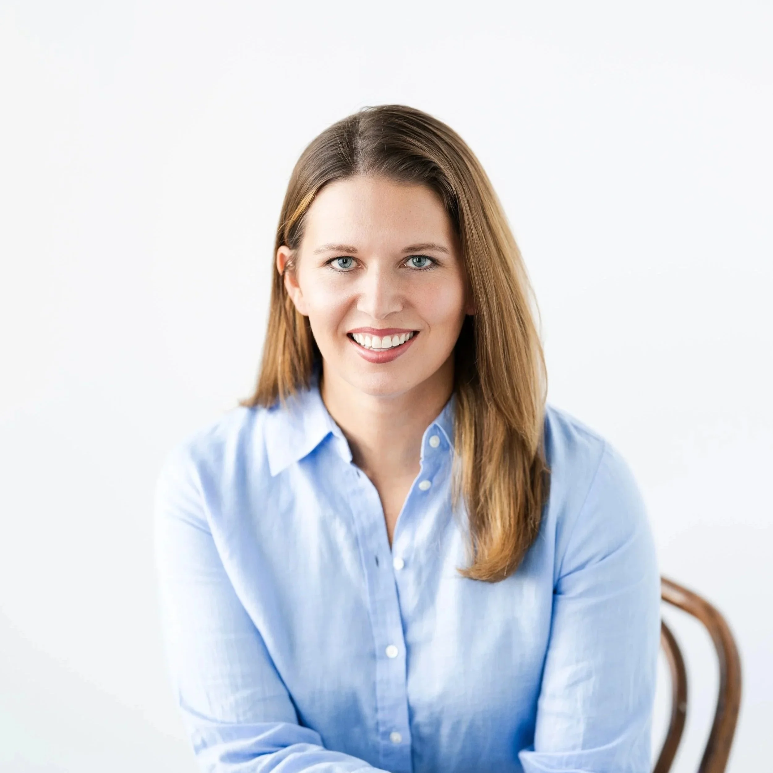 Lauren is pictured smiling wearing a blue button up shirt. Lauren has past shoulder length chestnut brown straight hair. Lauren is sitting on a brown chair with a white backgrounf.
