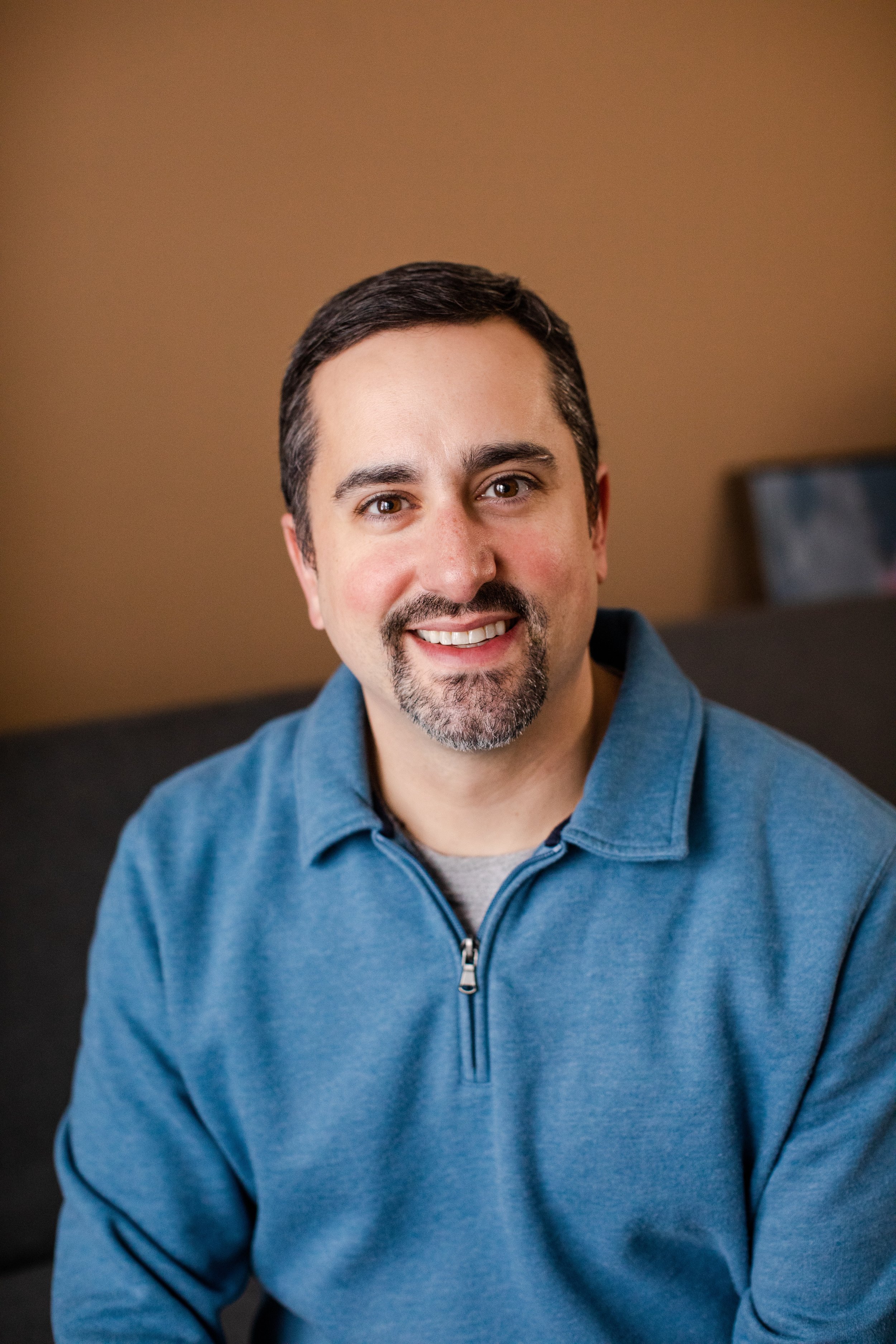 Noah is wearing a blue sweater with a color and zipper. He has dark hair and facial hair.