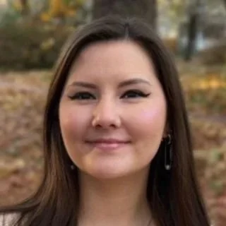 Chanel is wearing a beige sweater and has dark brown straight hair. Chanel is smiling closed mouth, has winged eyeliner and is wearing earrings. She is also standing in front of a blurred background of fallen leaves