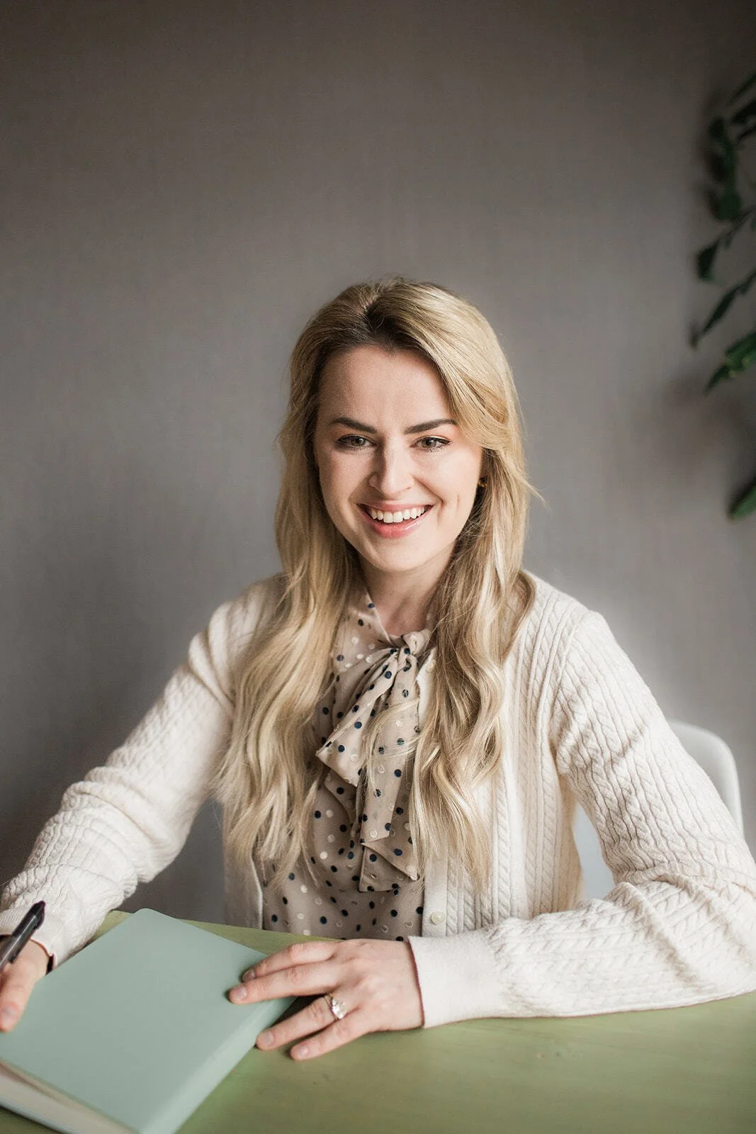 Sitting in front of a gray wall, Vera is smiling and has on light pink lipstick. Vera is wearing a beige knitted cardigan paired with a sand colored blouse that has black and white polka dots. Vera has chest length slightly wavy hair.