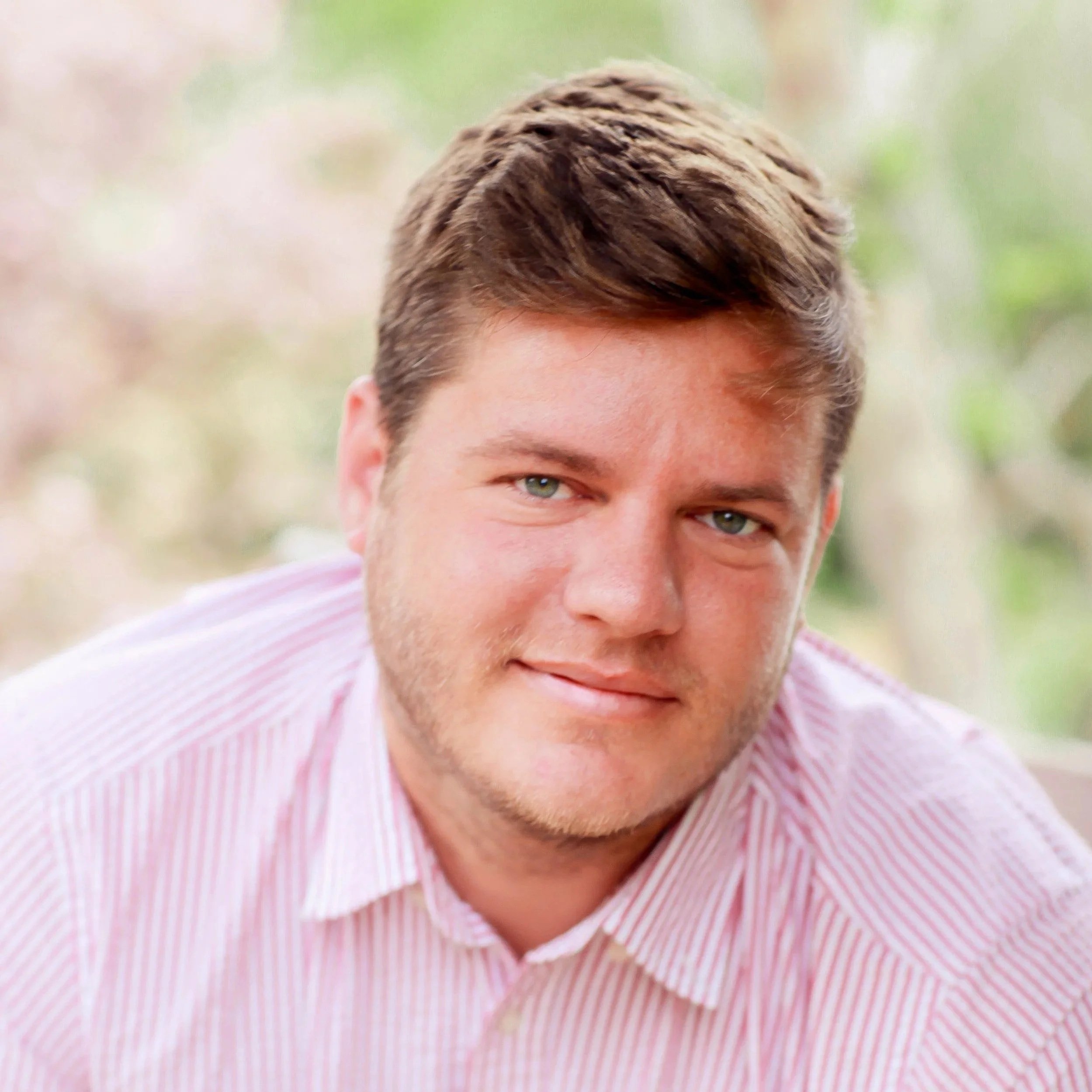 Andrew is pictured wearing a pink and white stripped button up. Andrew has light brown hair that is coiffed in the front.