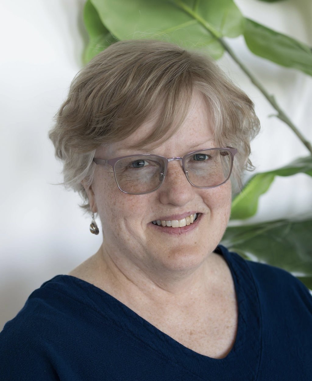 Karen is pictured smiling wearing a pink long sleeve and and blue lined glasses. Karen also has a pink multicolored scarf around her neck. Karen has an ear length wavy bob with brown and greying hair.