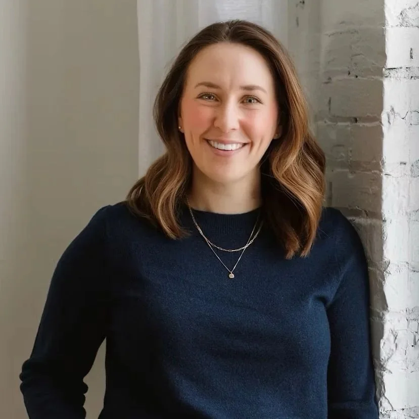 Mo is pictured smiling next to a white brick wall.  Mo has shoulder length caramel brown hair. Mo is wearing a navy blue blouse with a necklace.