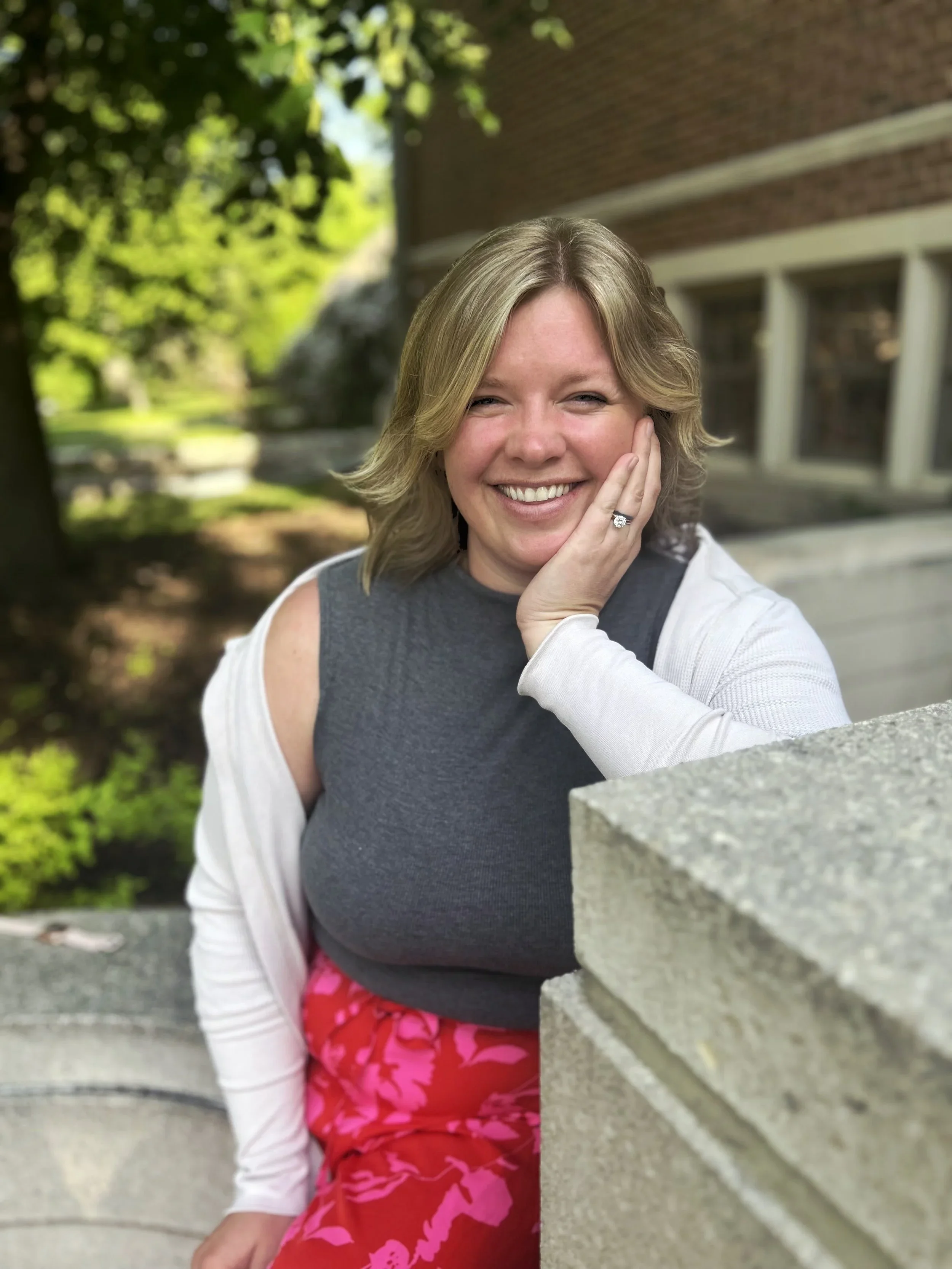 Sitting outside on a concrete bench, Abby is pictured smiling. Abby is wearing a dark grey tank top with a white cardigan as well as red pants with deep pink leaves printed on it. Abby has sandy blonde hair styled in a slightly wavy fashion.
