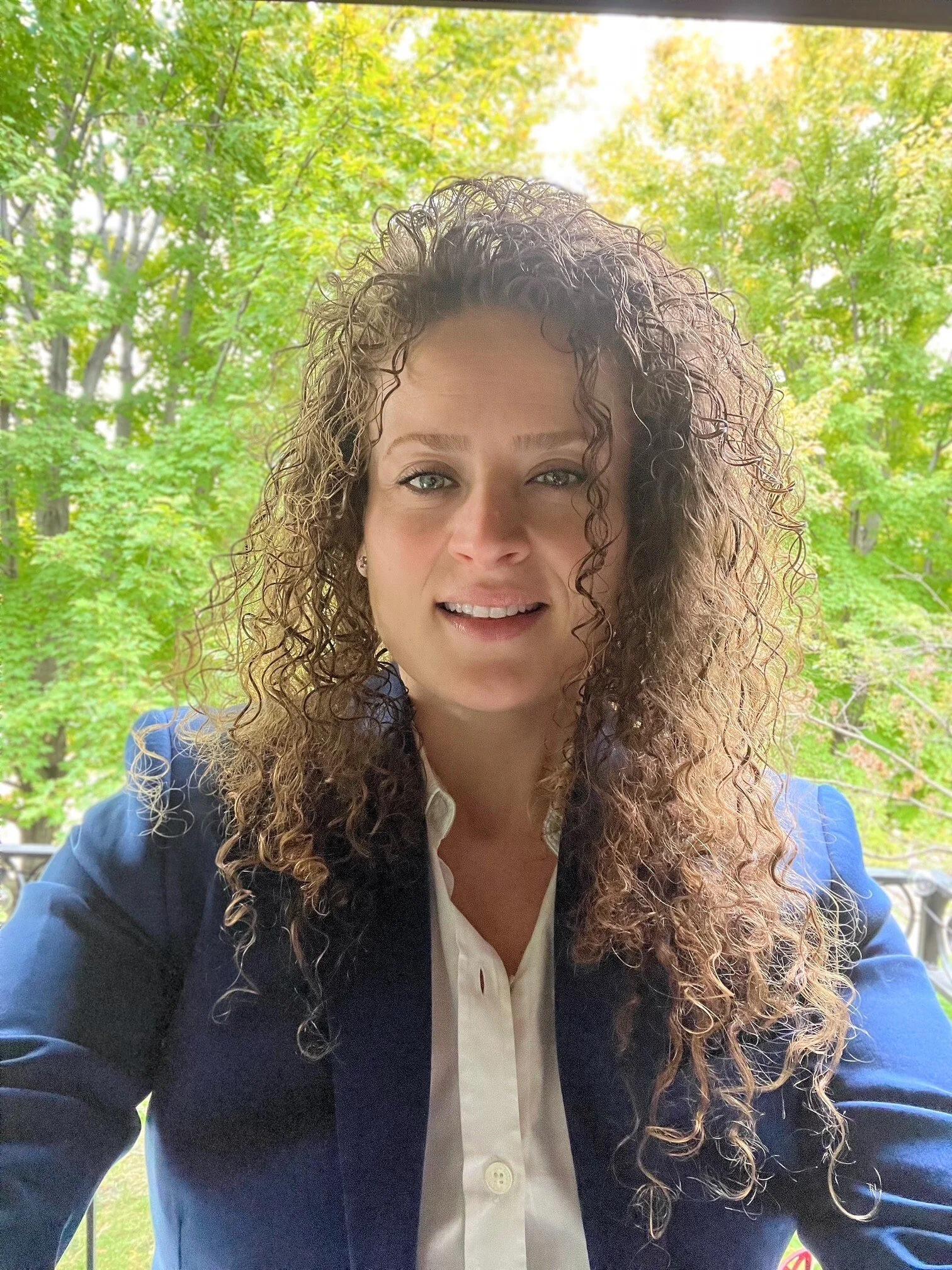 Mariya is pictured sitting outside with a background of trees and sunlight. Mariya has curly sandy blonde hair. Mariya is wearing a dark blue blaze with a white blouse.