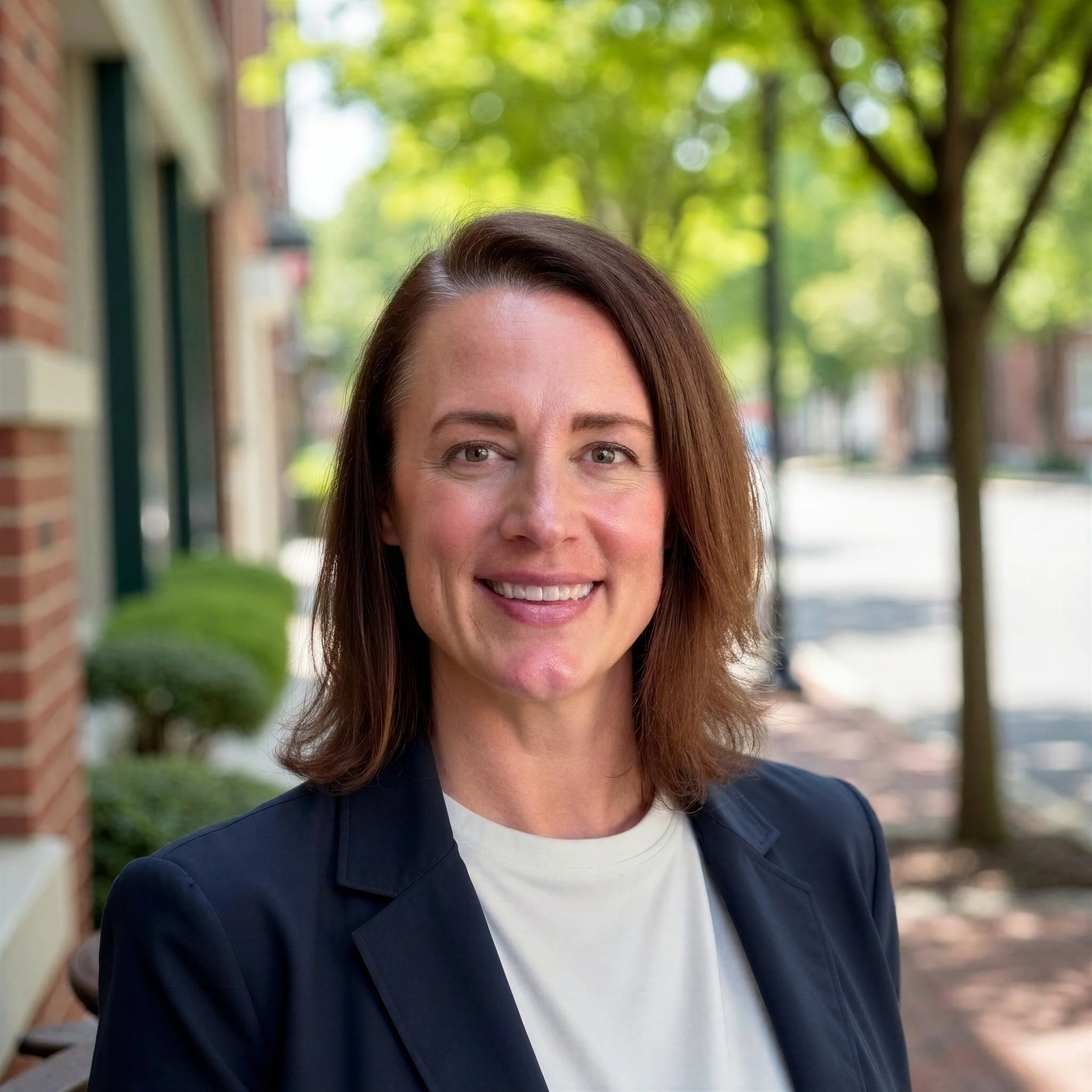 Dr. Kuhlmann is pictured smiling wearing a navy blue blazer with a white top underneath.  She has light brown should length hair. She is standing on a bricked pathway with trees in the background.