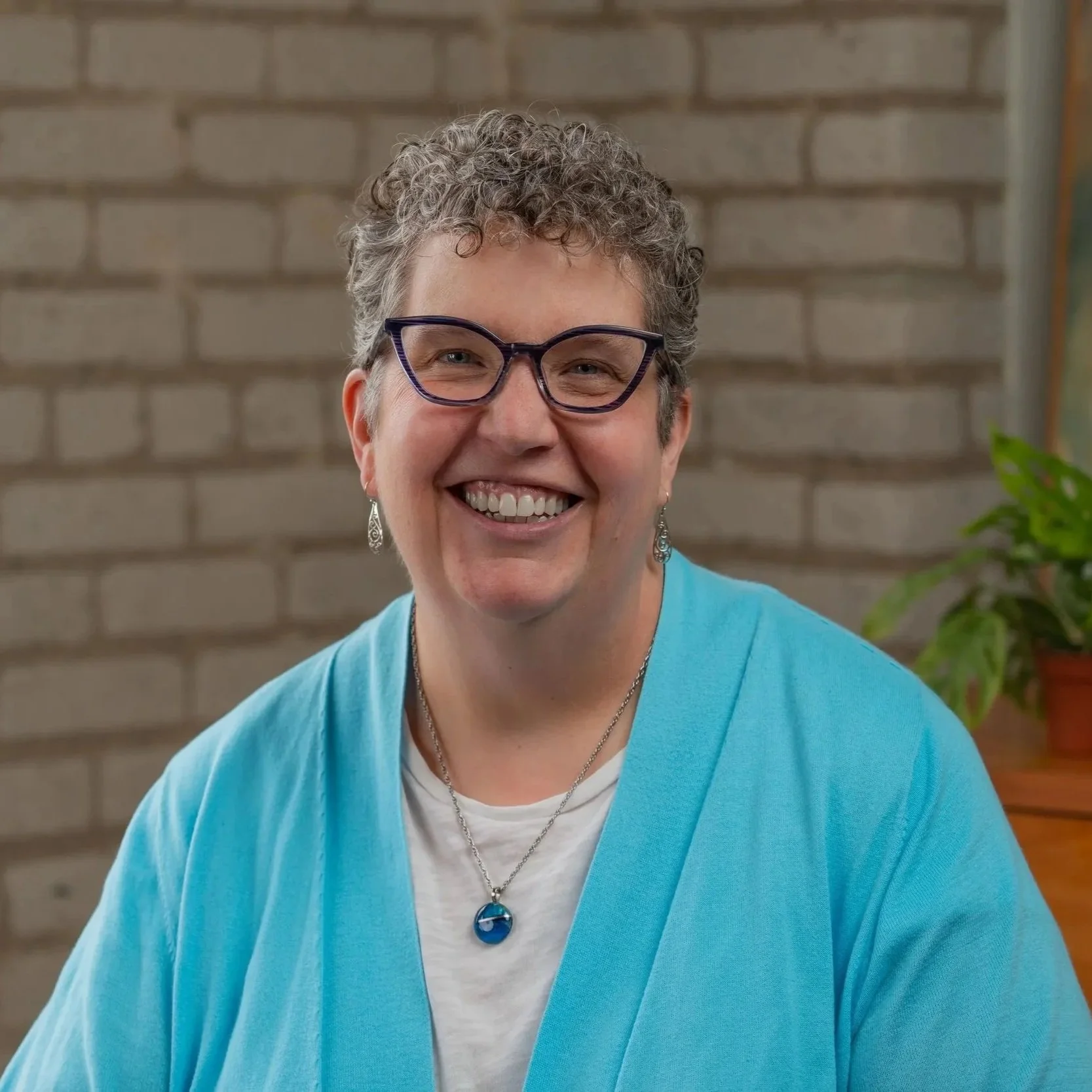 Susan is pictured smiling wearing a cyan blue cardigan with a white undershirt. Susan has salt and pepper colored hair with a low taper fade. Susan has on glasses, earrings and a blue stone necklace