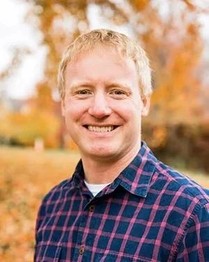 Karl is pictured smiling with a background of blurred leaves and trees during the fall. Karl has on a button up shirt with blue squares and pink lines. Karl has short light reddish hair.