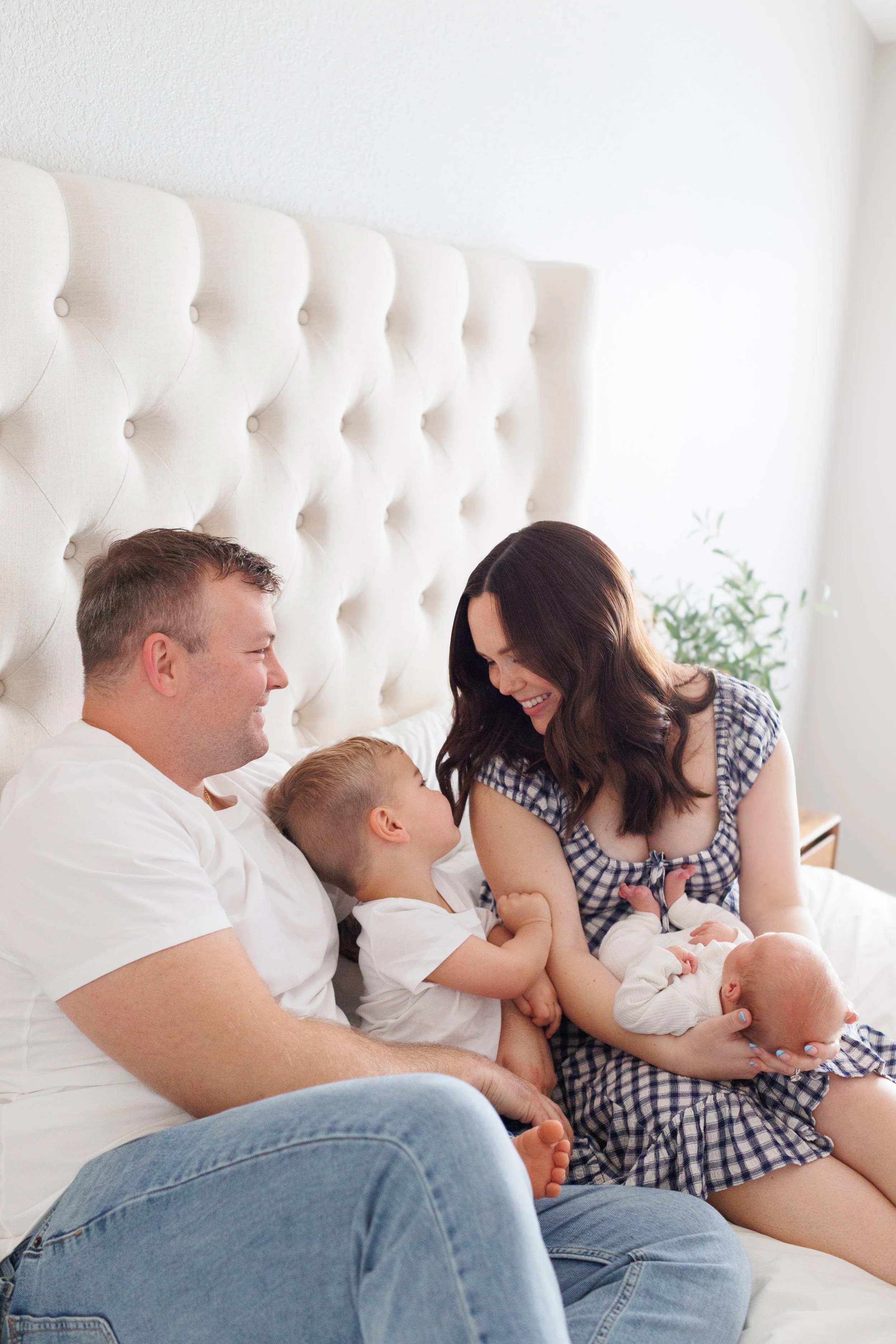 A happy family sitting on a bed, including a father, mother, a young child, and a baby, all smiling at each other.