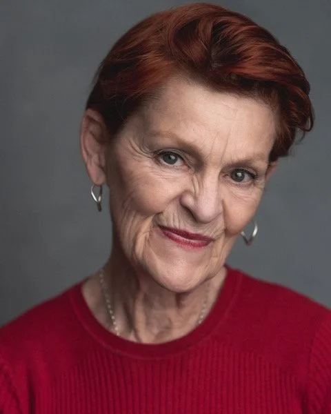 Close-up portrait of a smiling elderly woman with short, red hair, wearing a red top and hoop earrings, against a gray background.